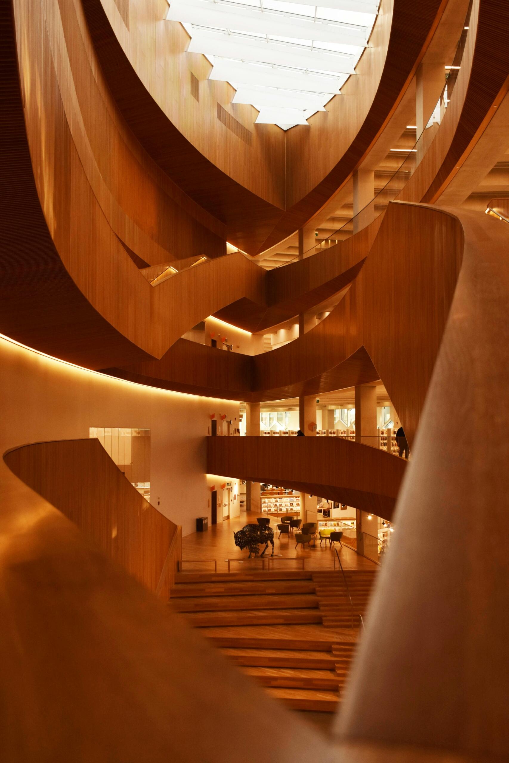 A photo of a modern atrium and staircase. The entire space is made of light wood. A warm light illuminates the space. Source: Nikita.