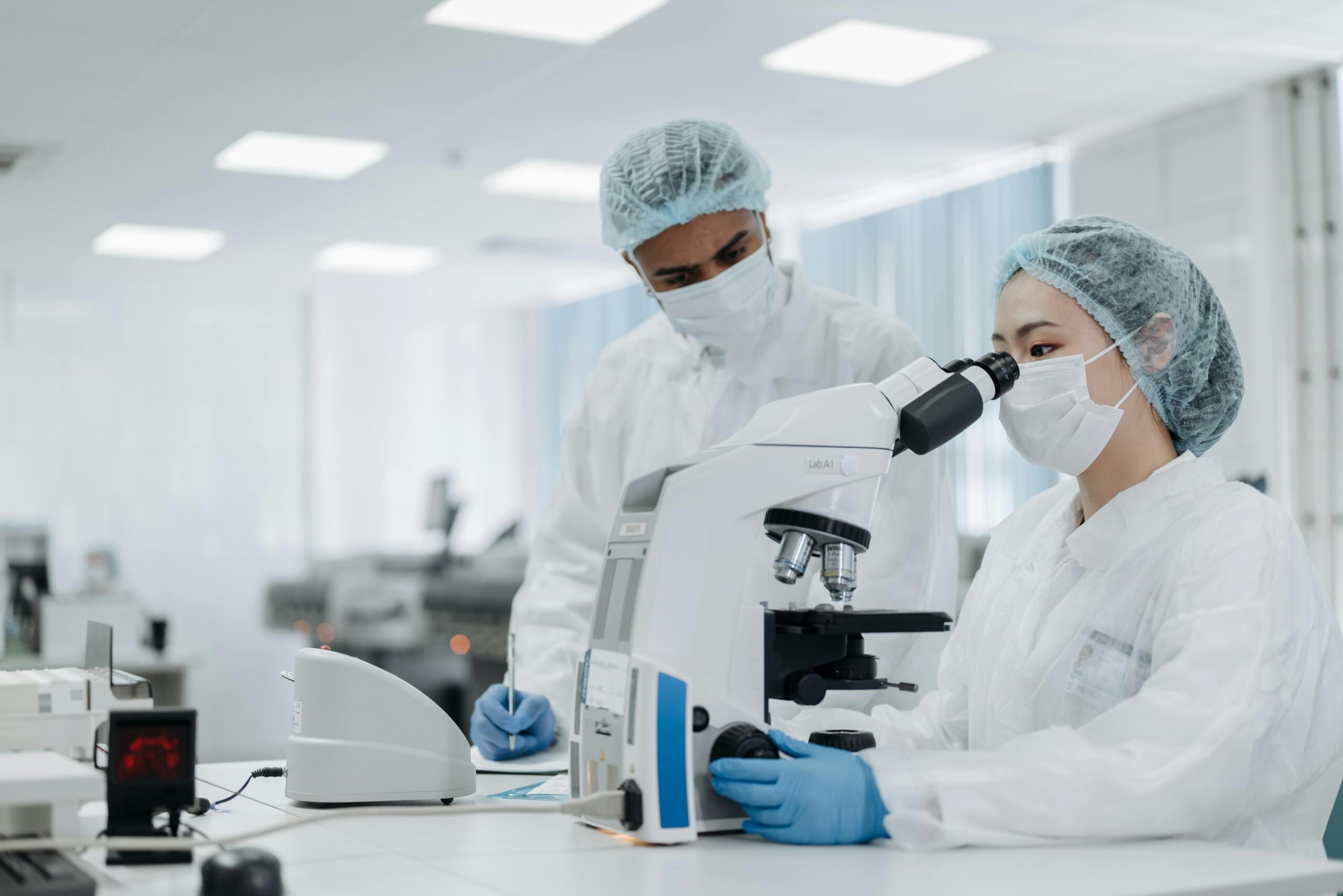 Two researchers in full protective gear analyzing samples using a microscope in a modern laboratory.