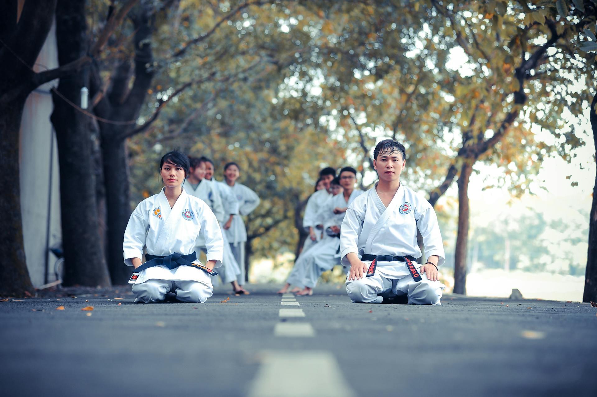 People in karate uniform on their knees at the park surrounded by trees.
