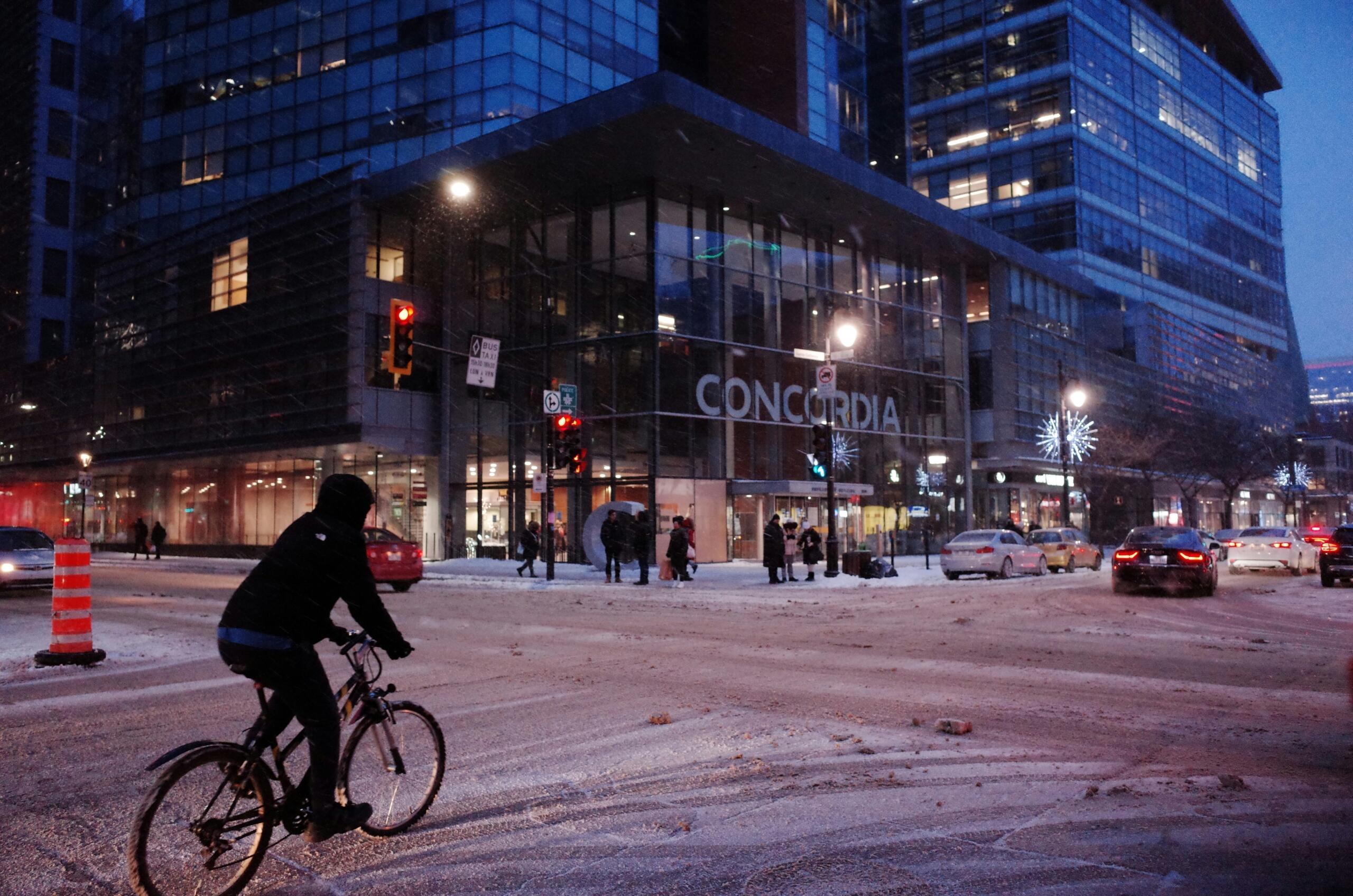 A winter evening scene featuring Concordia University’s modern glass building in Montreal. A cyclist rides past while pedestrians walk along the snow-covered street.