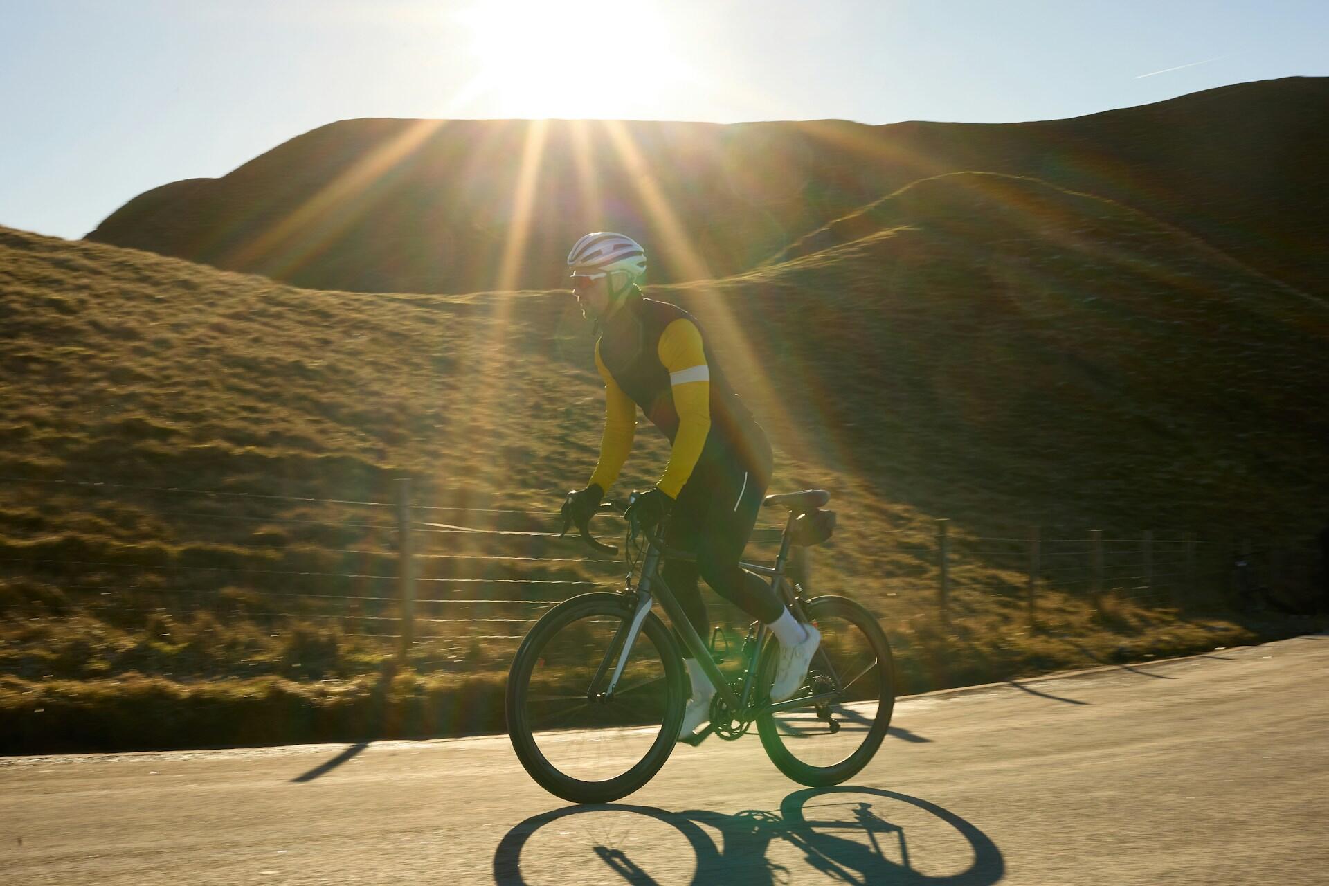 Cyclist riding on a road with hill background, sun shining from behind.