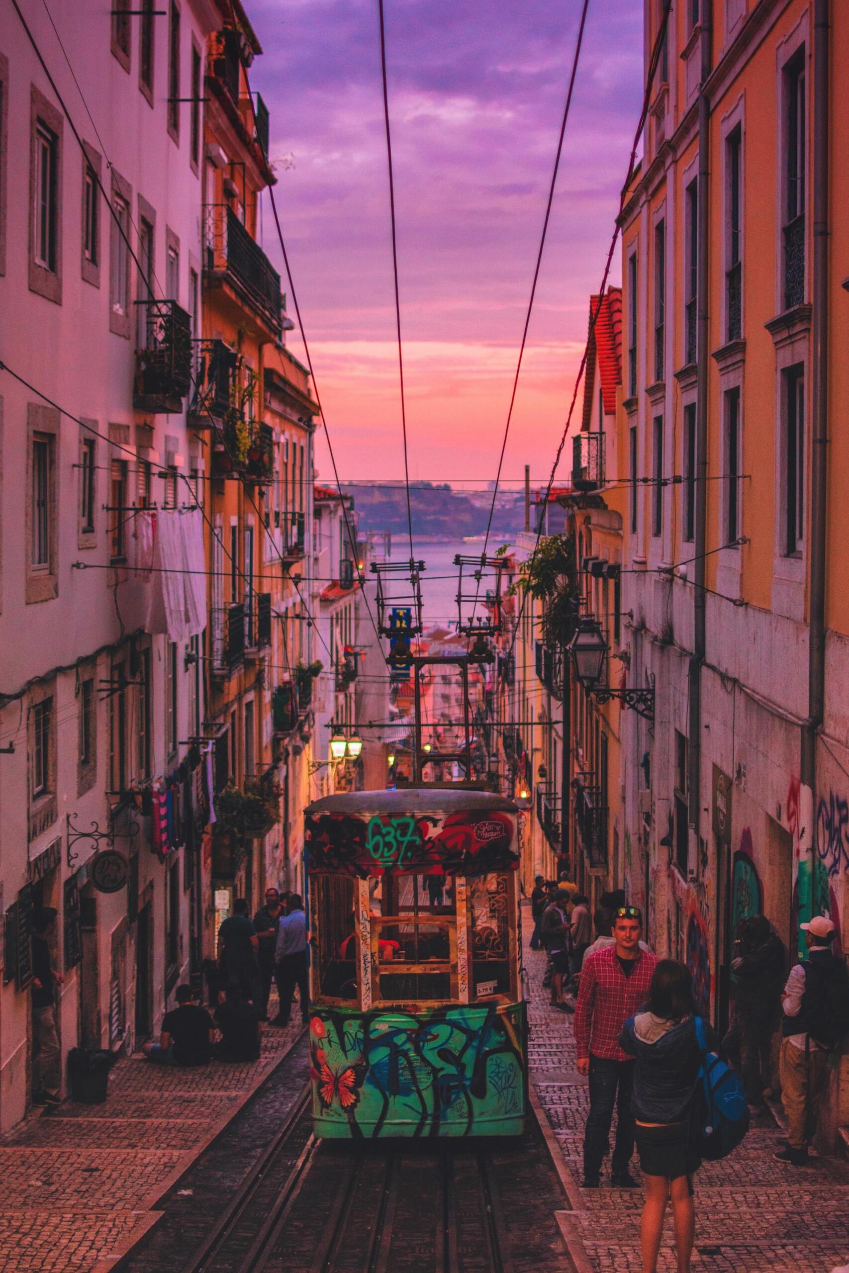 A vibrant street scene captures a colorful tram descending steep cobblestones, framed by pastel buildings under a stunning sunset sky.