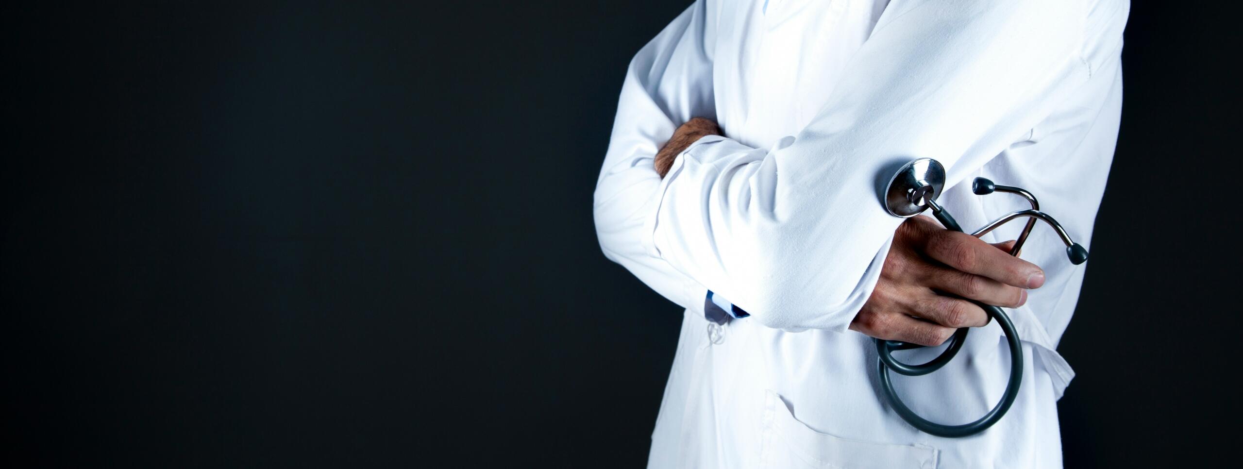 A close-up of a doctor wearing a white coat and holding a stethoscope, standing with arms crossed against a dark background.