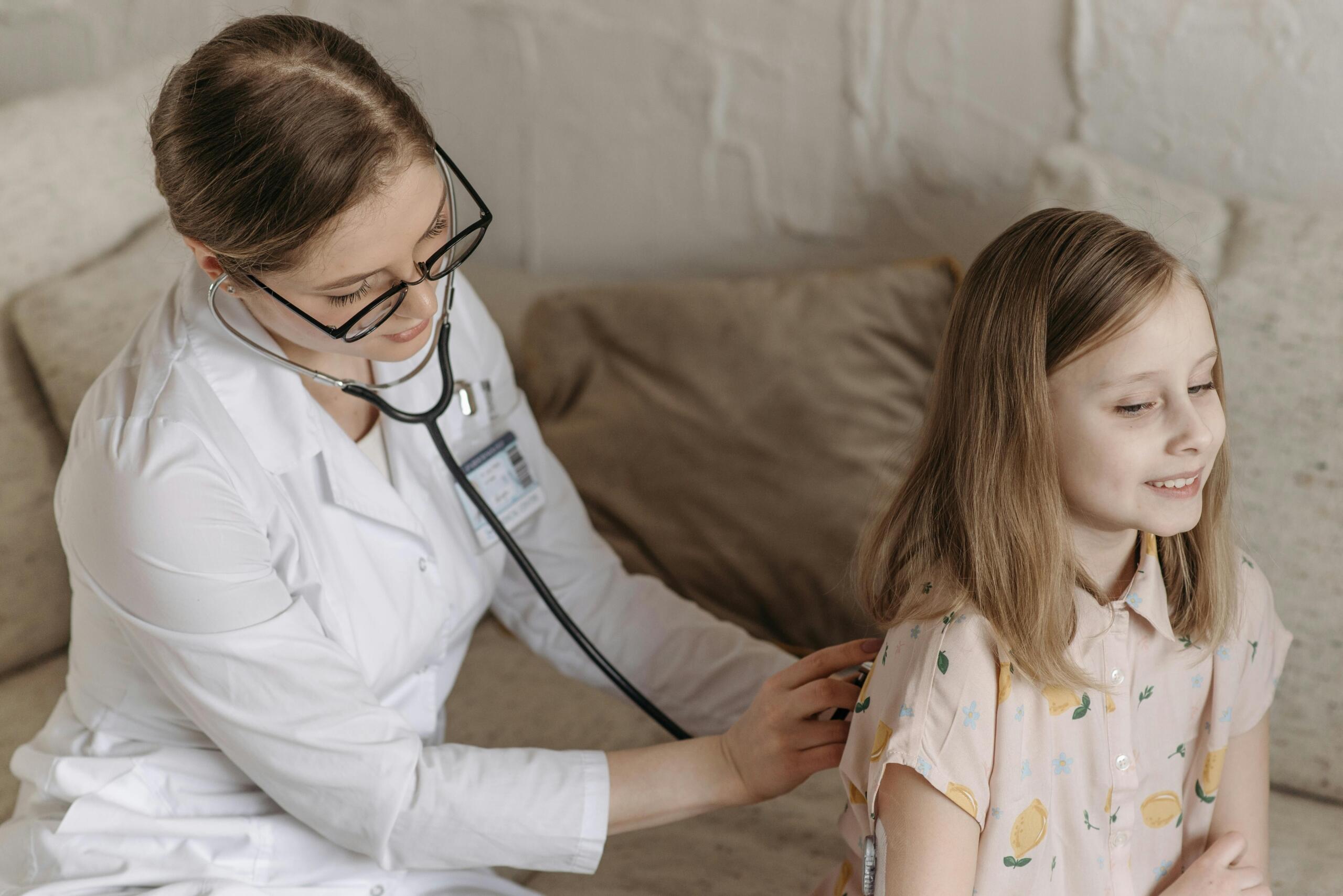 A female doctor using a stethoscope to examine a young girl sitting on a couch, symbolizing patient care in pediatrics.