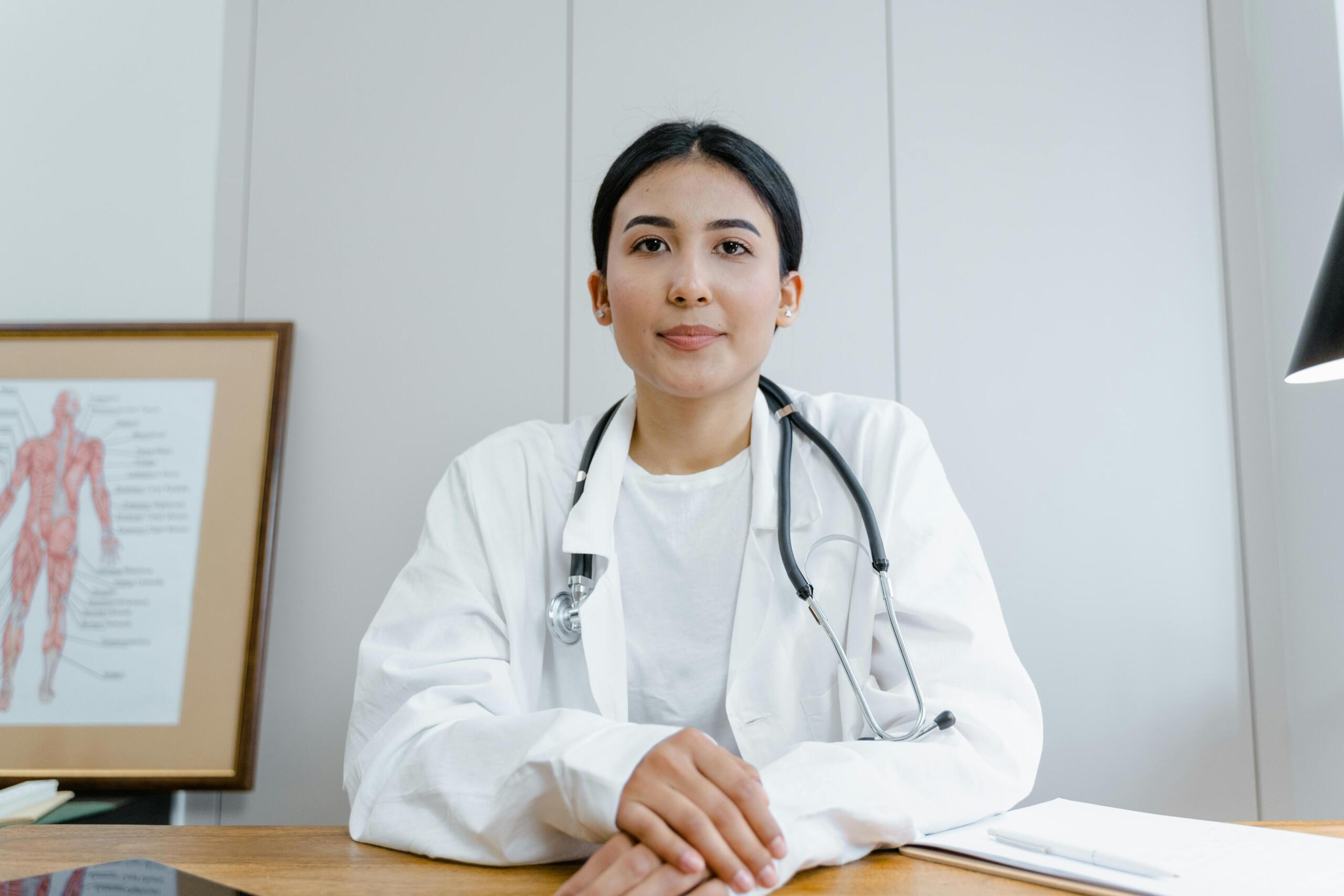 A young doctor in a white coat sitting at a desk with a stethoscope around her neck, ready for consultation.