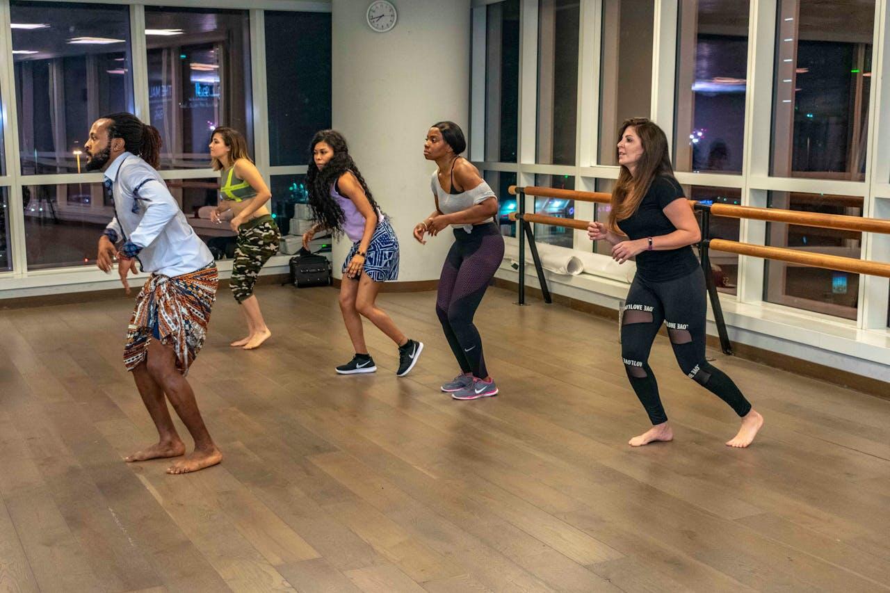 A dance class with five participants performing synchronized movements in a brightly lit studio with large windows.