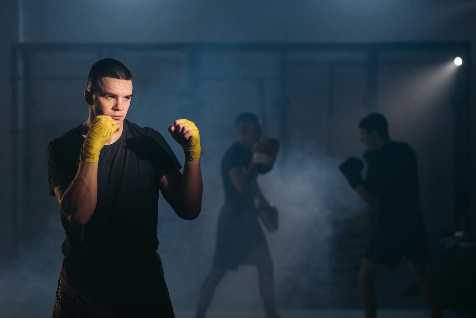 A Man in Black Shirt Wearing Yellow Hand Wraps, a boxer is hitting pads in the background, clouded by smoke.