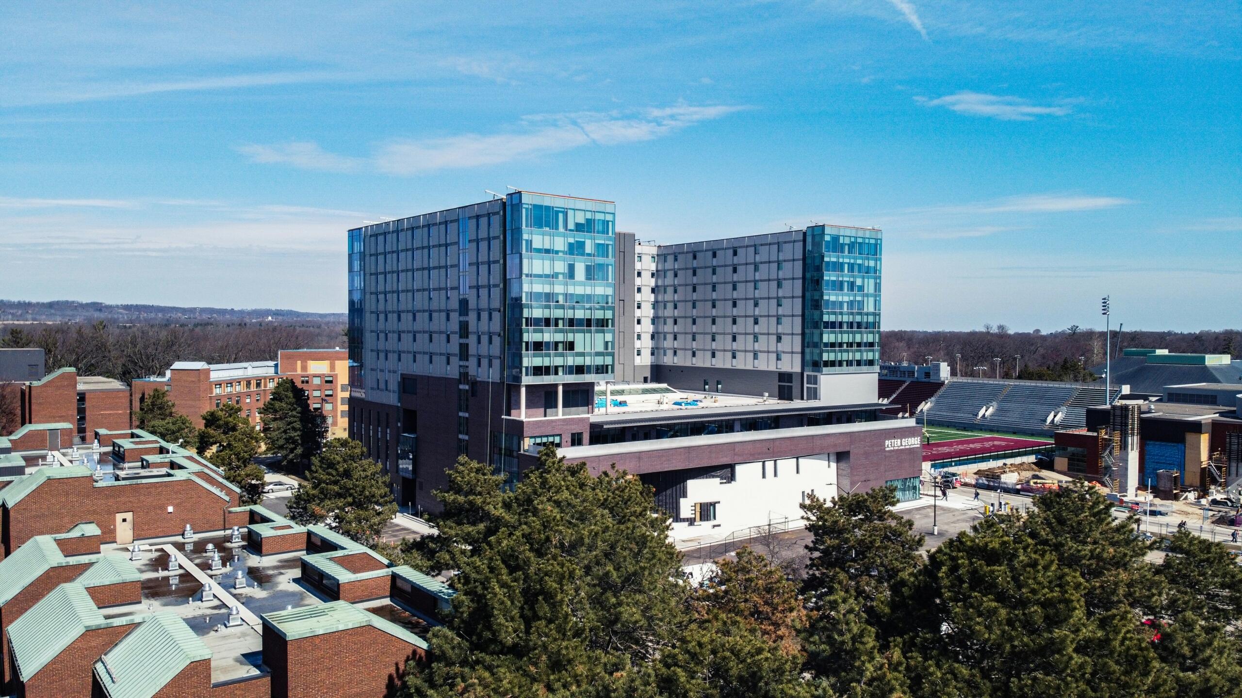 A large, modern multi-story building on the McMaster University campus, featuring glass windows reflecting the bright blue sky. Surrounding the building are older brick structures and green trees, with a stadium visible in the background.