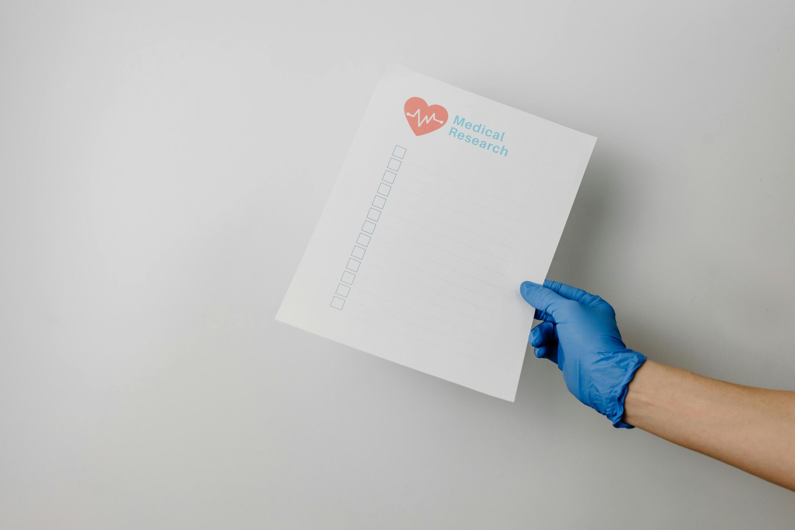 A gloved hand holding a medical research checklist with a heart logo at the top, representing organized research documentation.