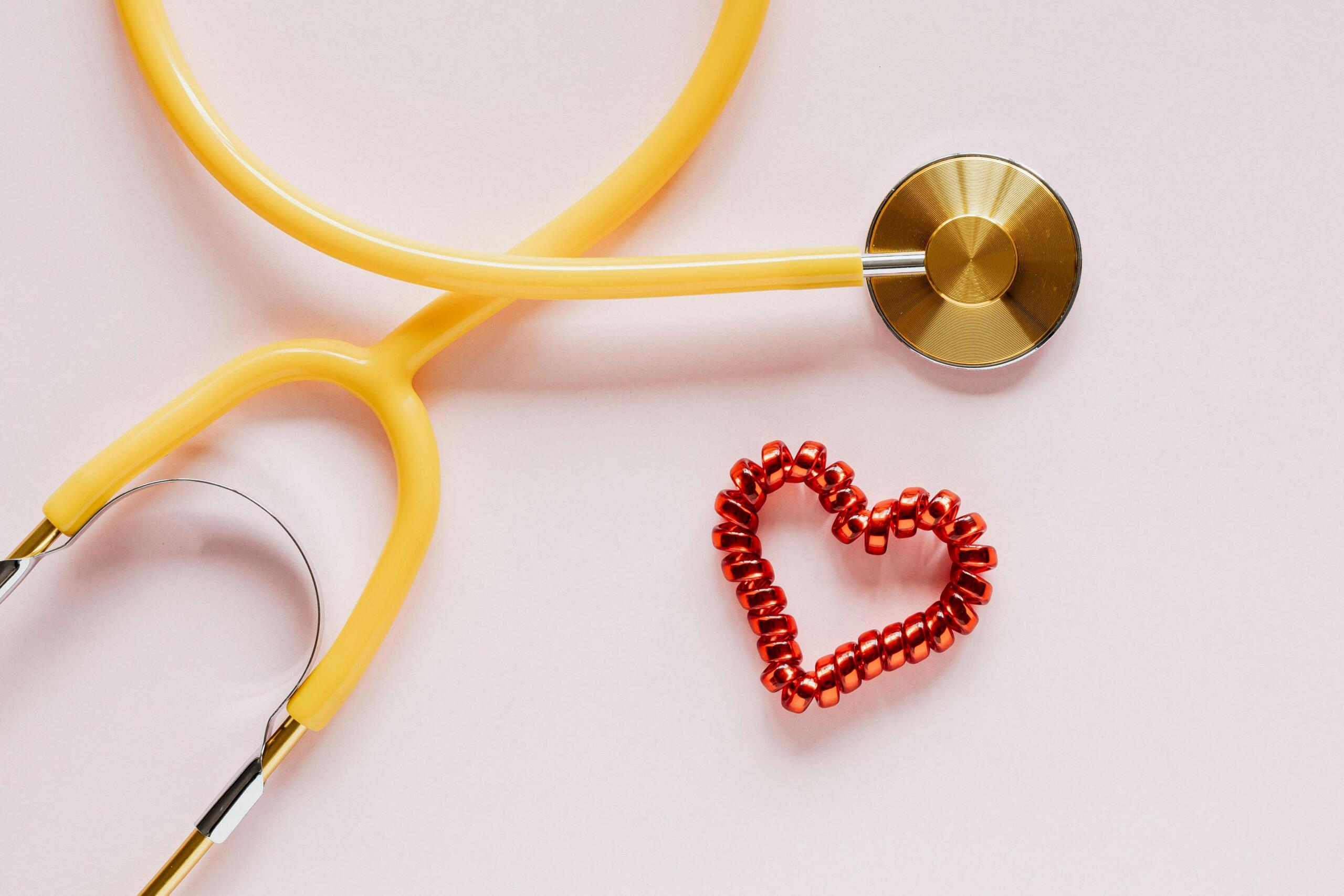 A yellow stethoscope placed next to a red heart-shaped decoration on a light pink background, representing healthcare and compassion in the medical field.