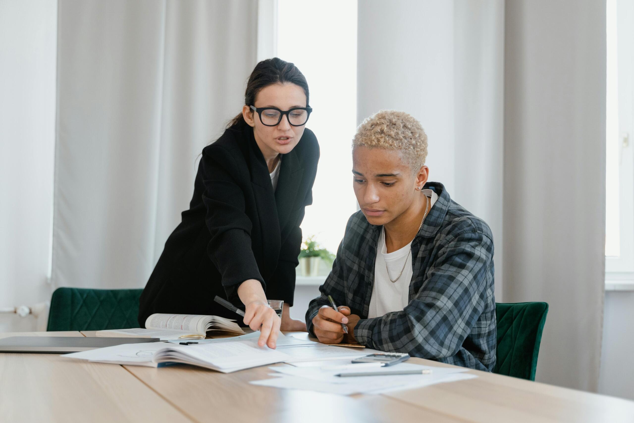 A student and a tutor working together at a table with study materials. The tutor, wearing glasses and a blazer, is pointing at a document while the student attentively listens and takes notes.
