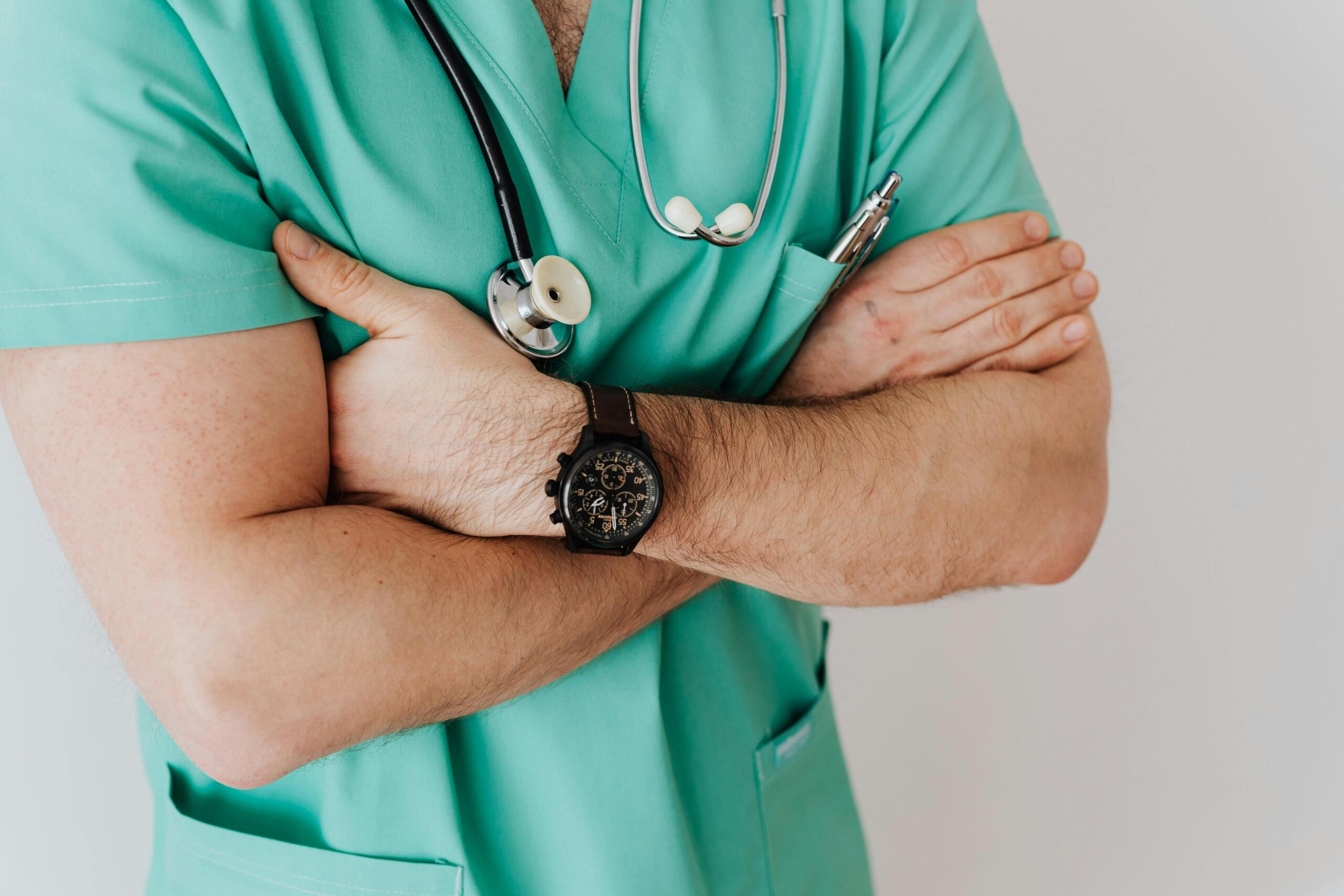 A male healthcare professional in green scrubs, wearing a stethoscope around his neck and crossing his arms.