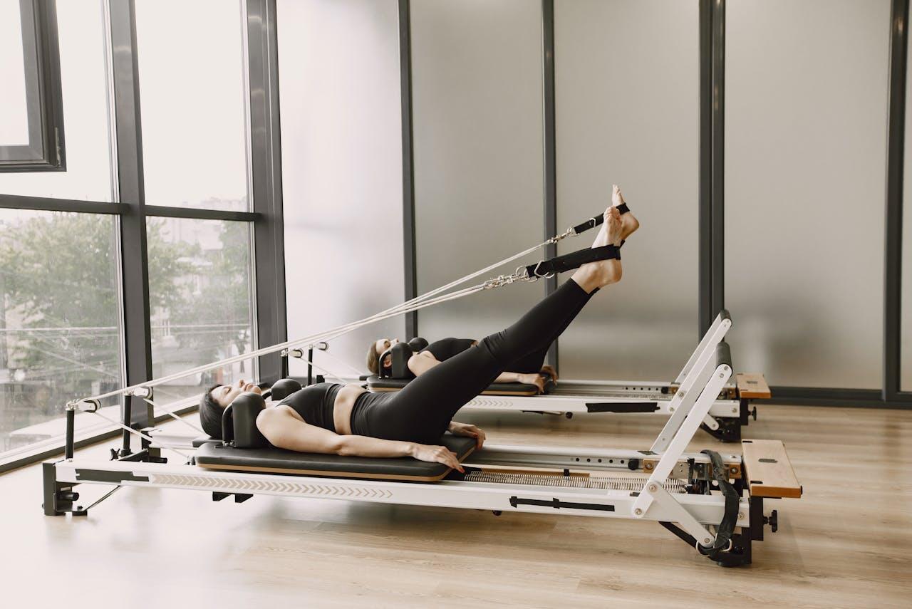 A person performs Pilates exercises on a reformer, with legs extended and straps attached, in a bright, airy studio.