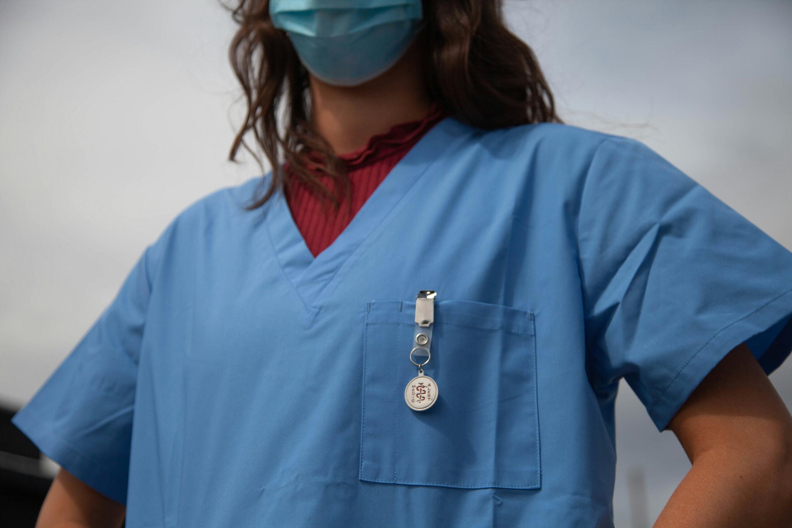 A healthcare professional in blue scrubs with an ID badge, representing postgraduate medical training in a hospital environment.