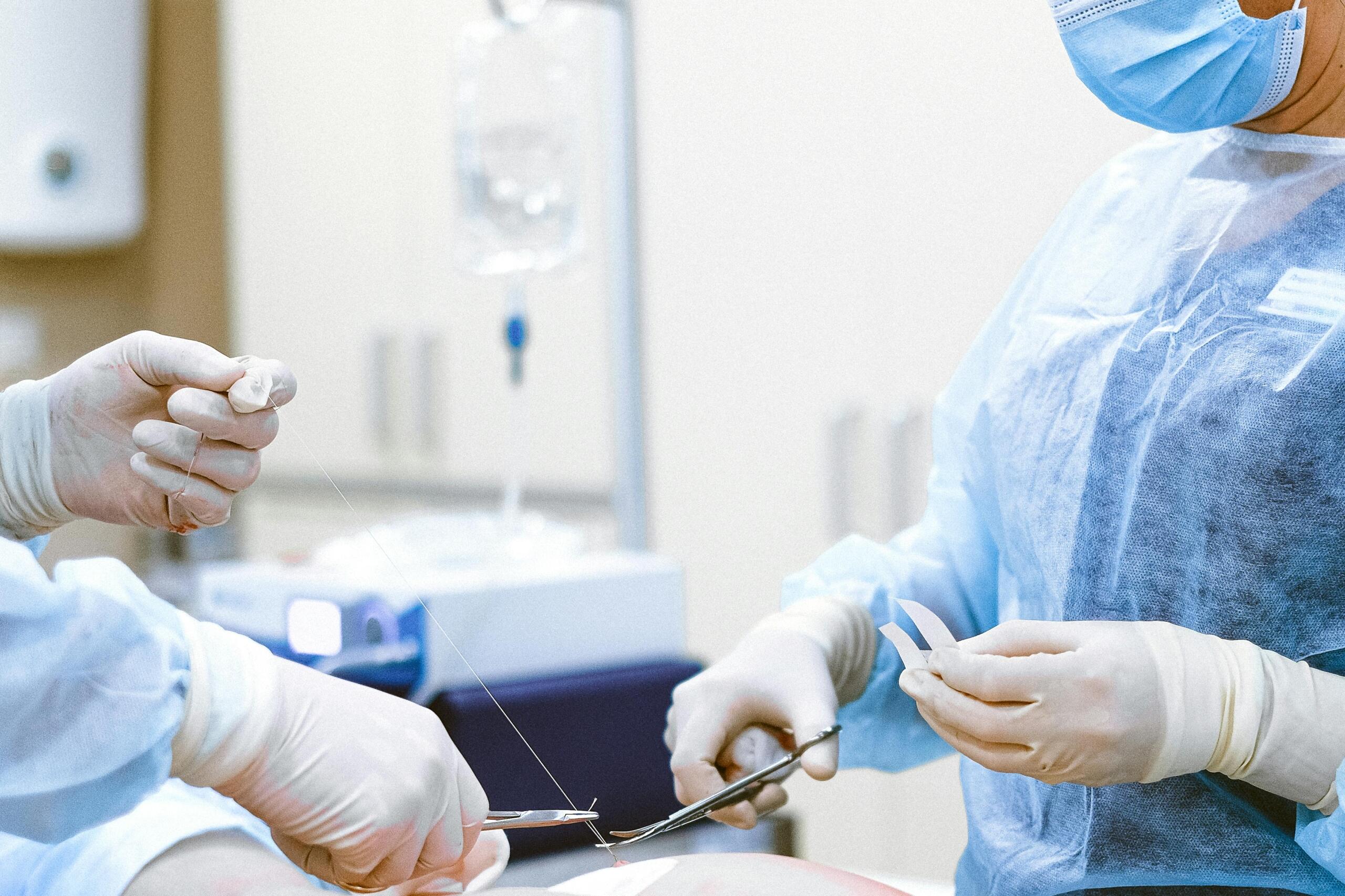 Two surgeons wearing surgical gowns and gloves performing a procedure, one holding a surgical instrument and thread for suturing.