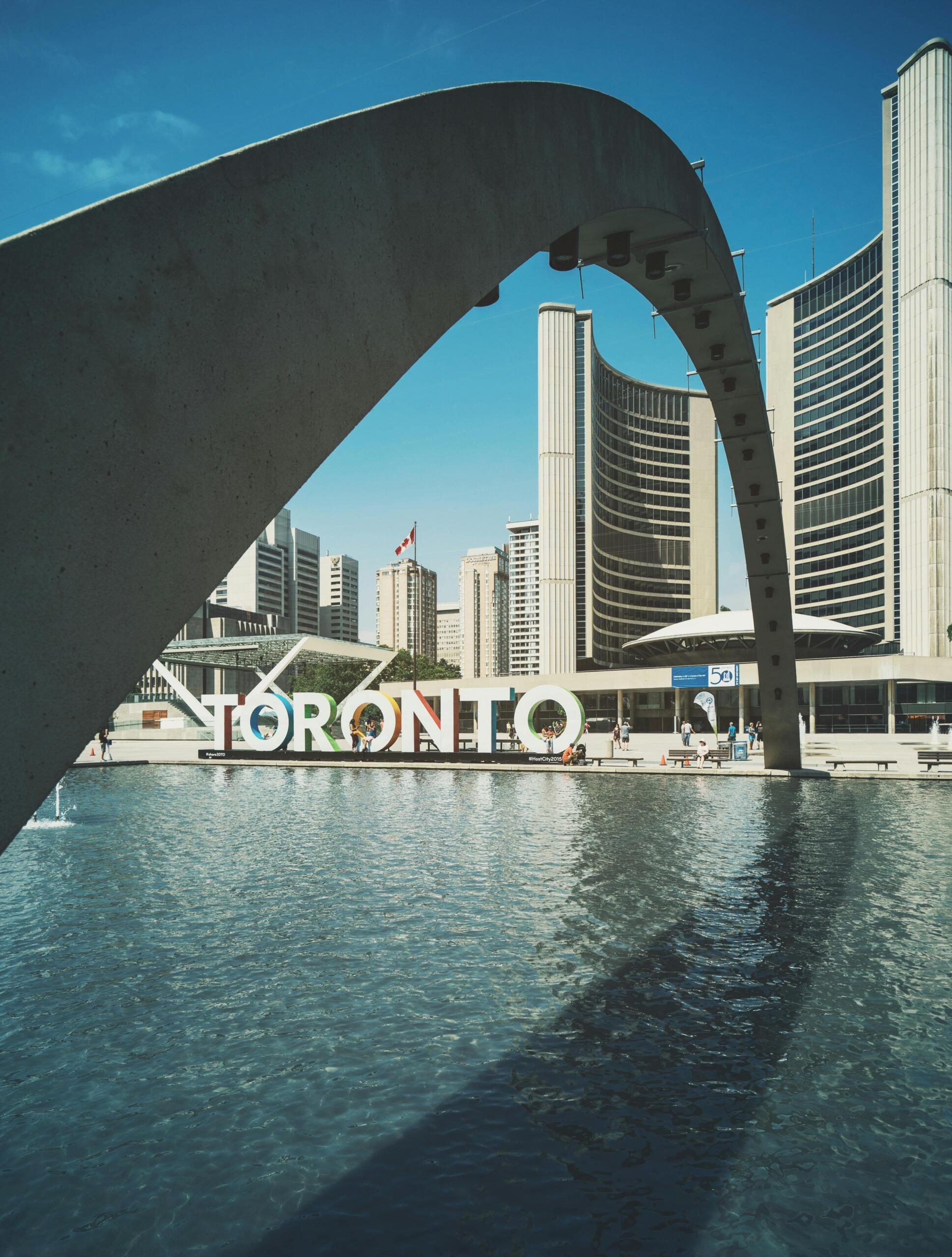 Two tall curved buildings are pictured behind a sign that says "Toronto". A large water fountain and a curved structure are in front of the curved buildings.