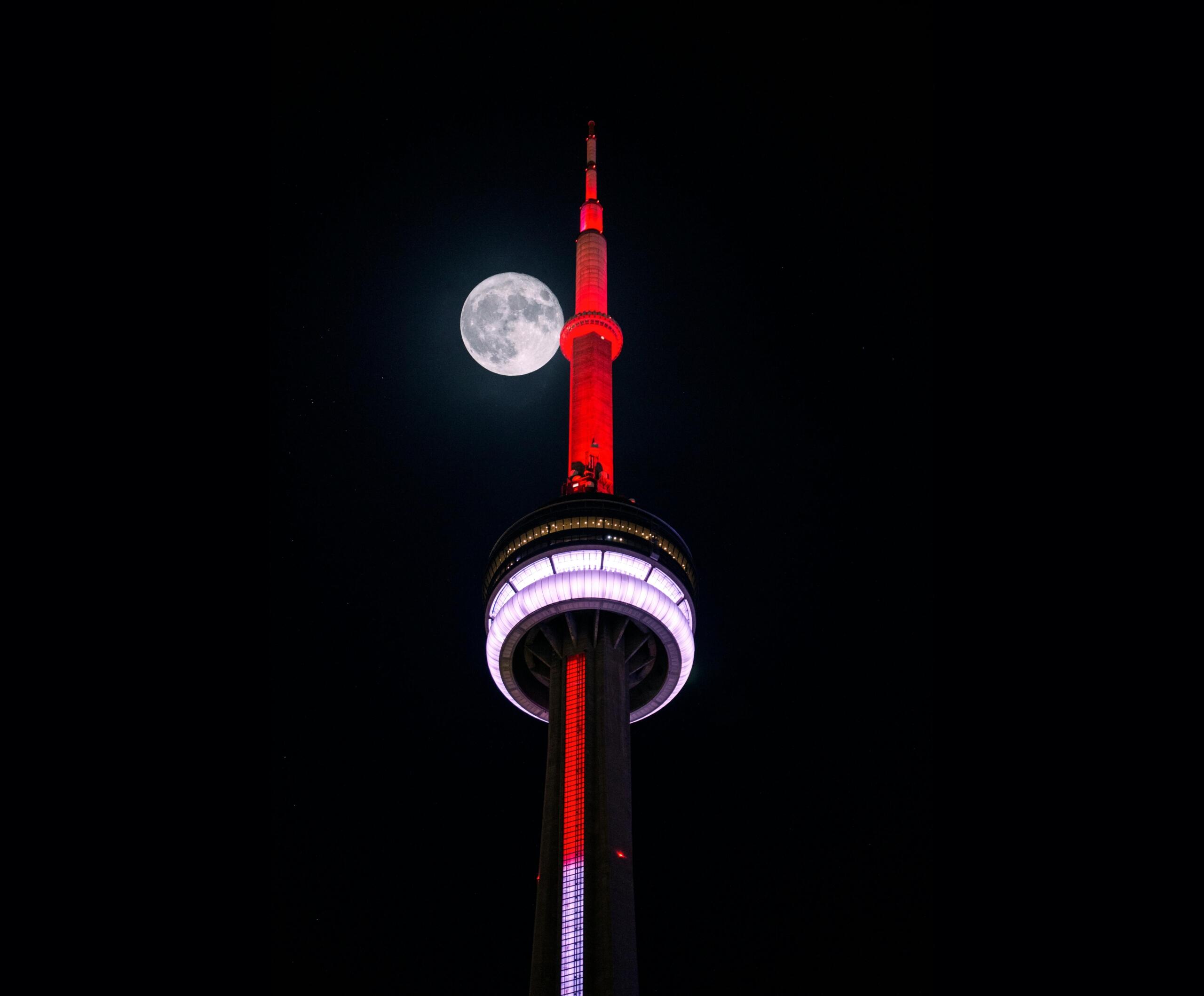 Toronto's CN tower at night. The tower is light up in red and a full moon can be seen behind the building.