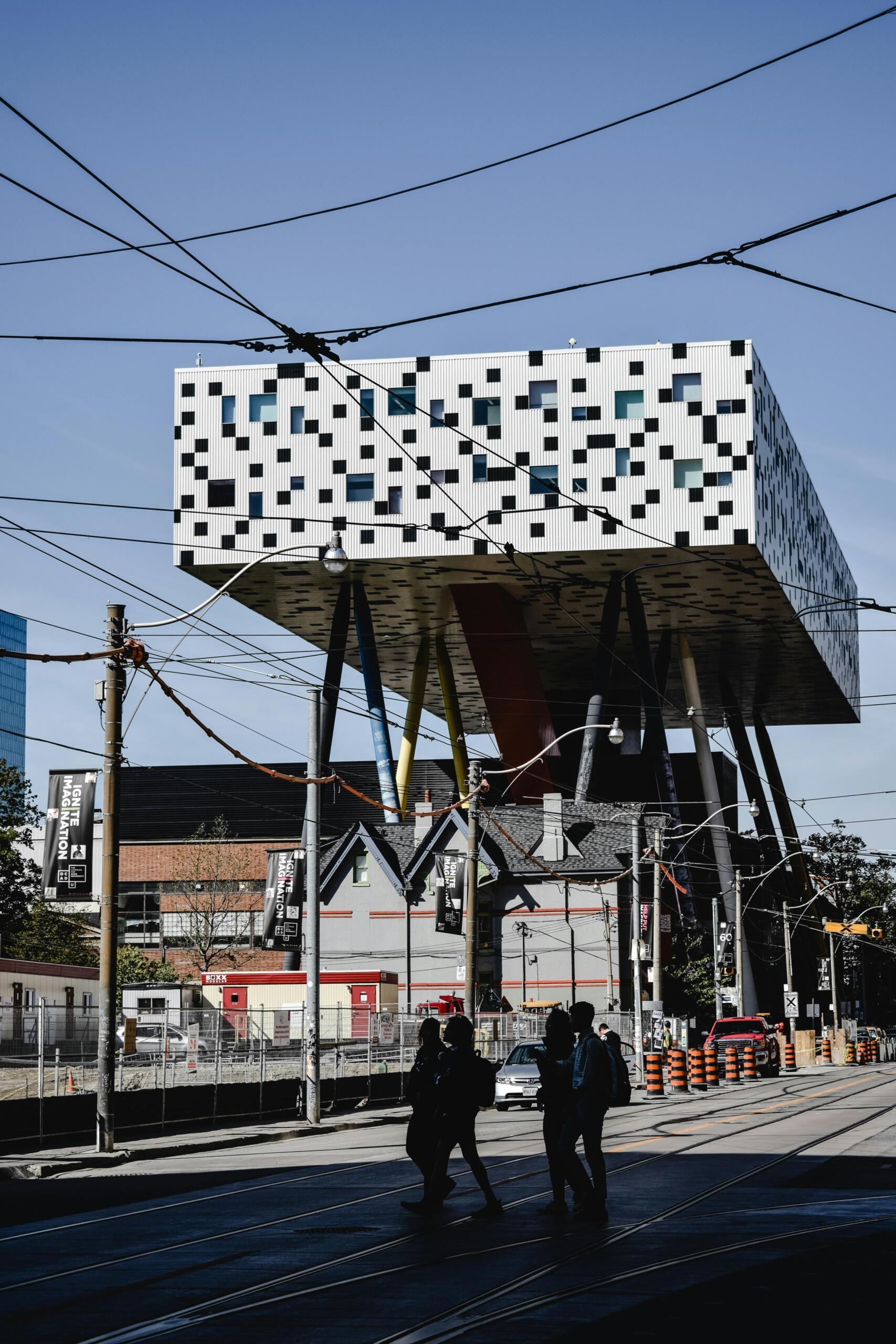 A white building with blue squares on a city street. The building sits atop long tubes.