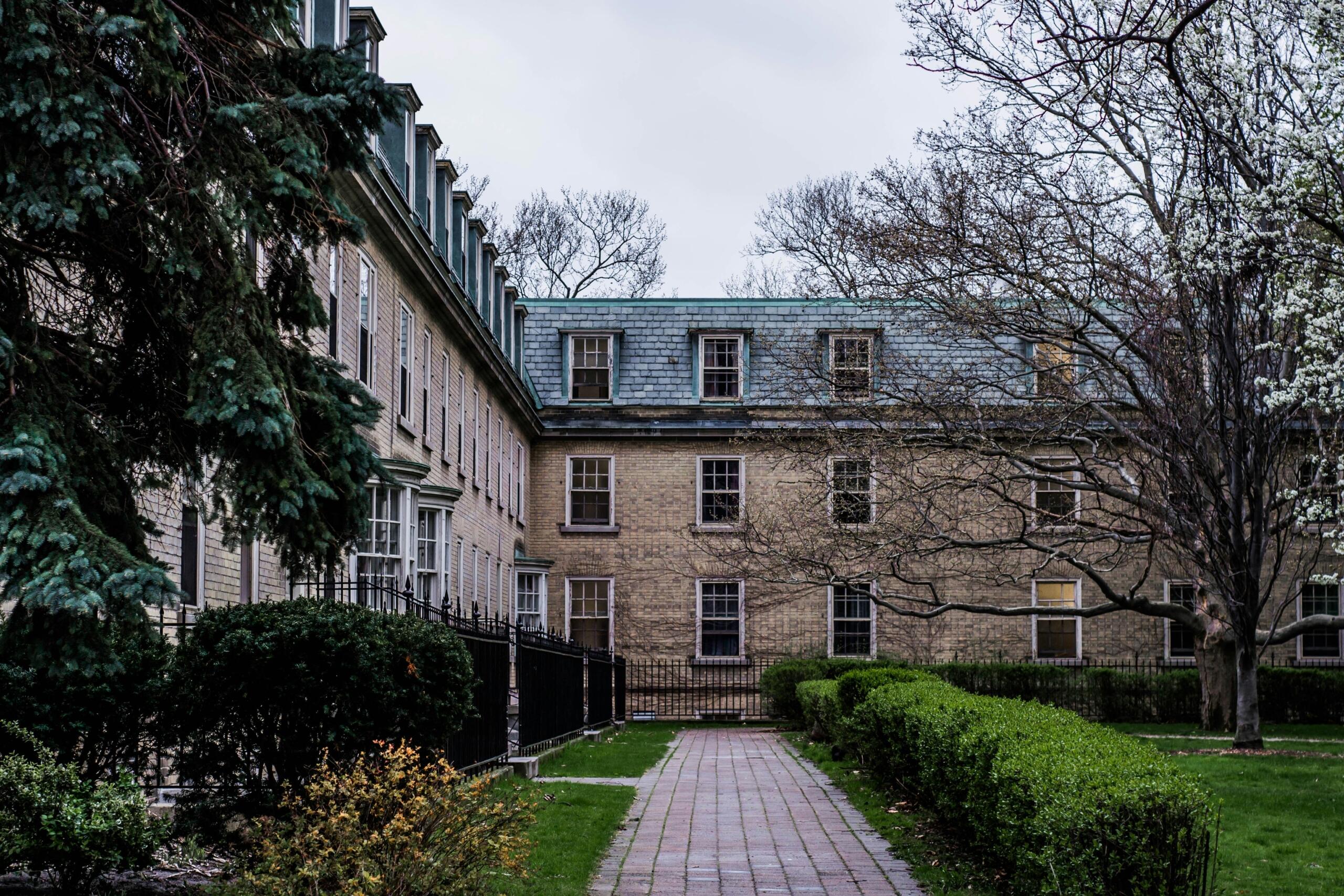 A historic brick building on the University of Toronto St. George campus, with a tree-lined pathway leading to the entrance. The setting is peaceful, with neatly trimmed hedges and a cloudy sky above.