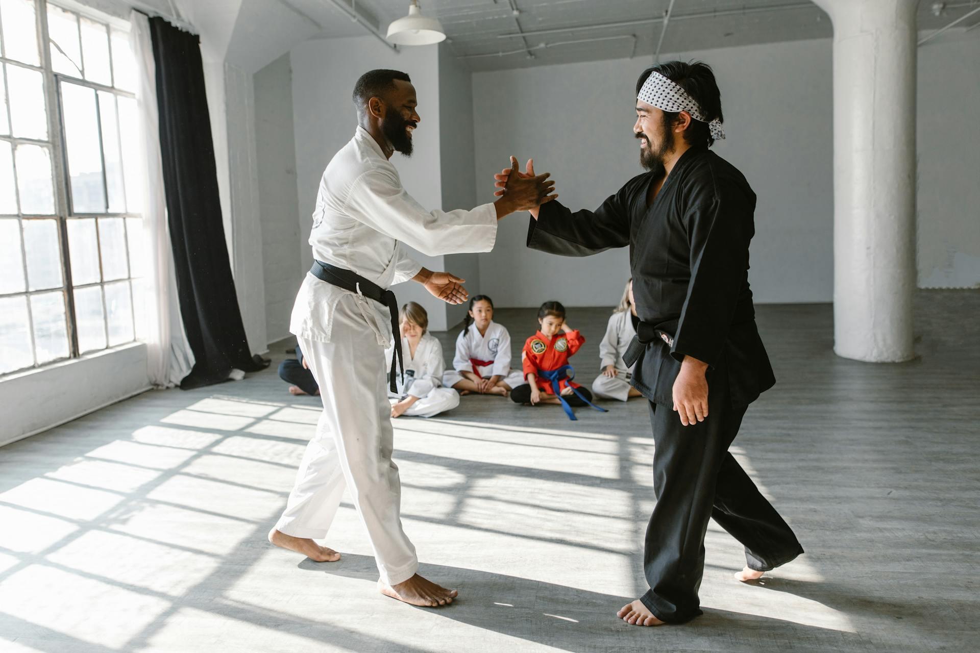 We see two MA coaches greeting each other, one wearing a black gi and black belt, one wearing a white gi and black belt. Students look on.