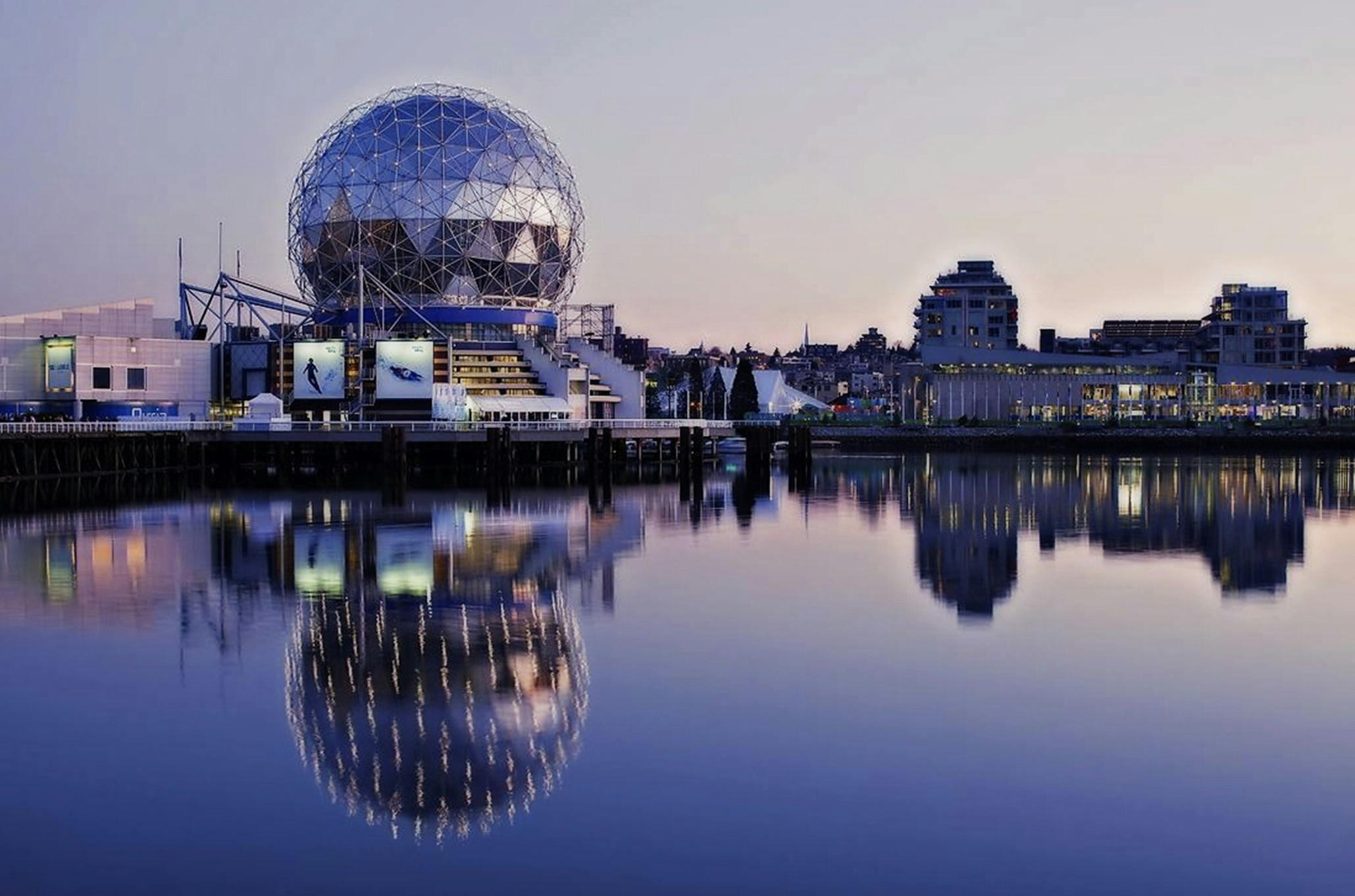 A geodesic dome on a waterfront location is shown at dusk. The dome reflects into the water.