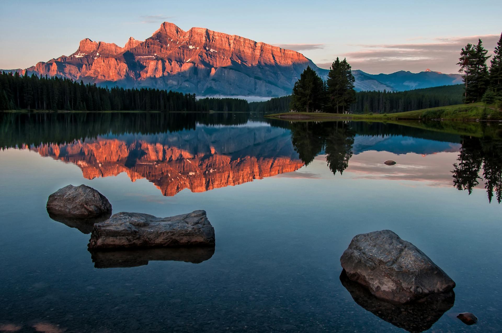 Majestic mountains reflect in a calm lake, with rocky foreground and lush trees lining the shore under a serene sky.