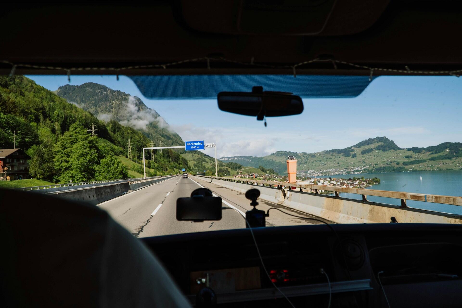 View from a vehicle driving on a highway, with mountains and a lake in the background, approaching a "Beckenried" sign.