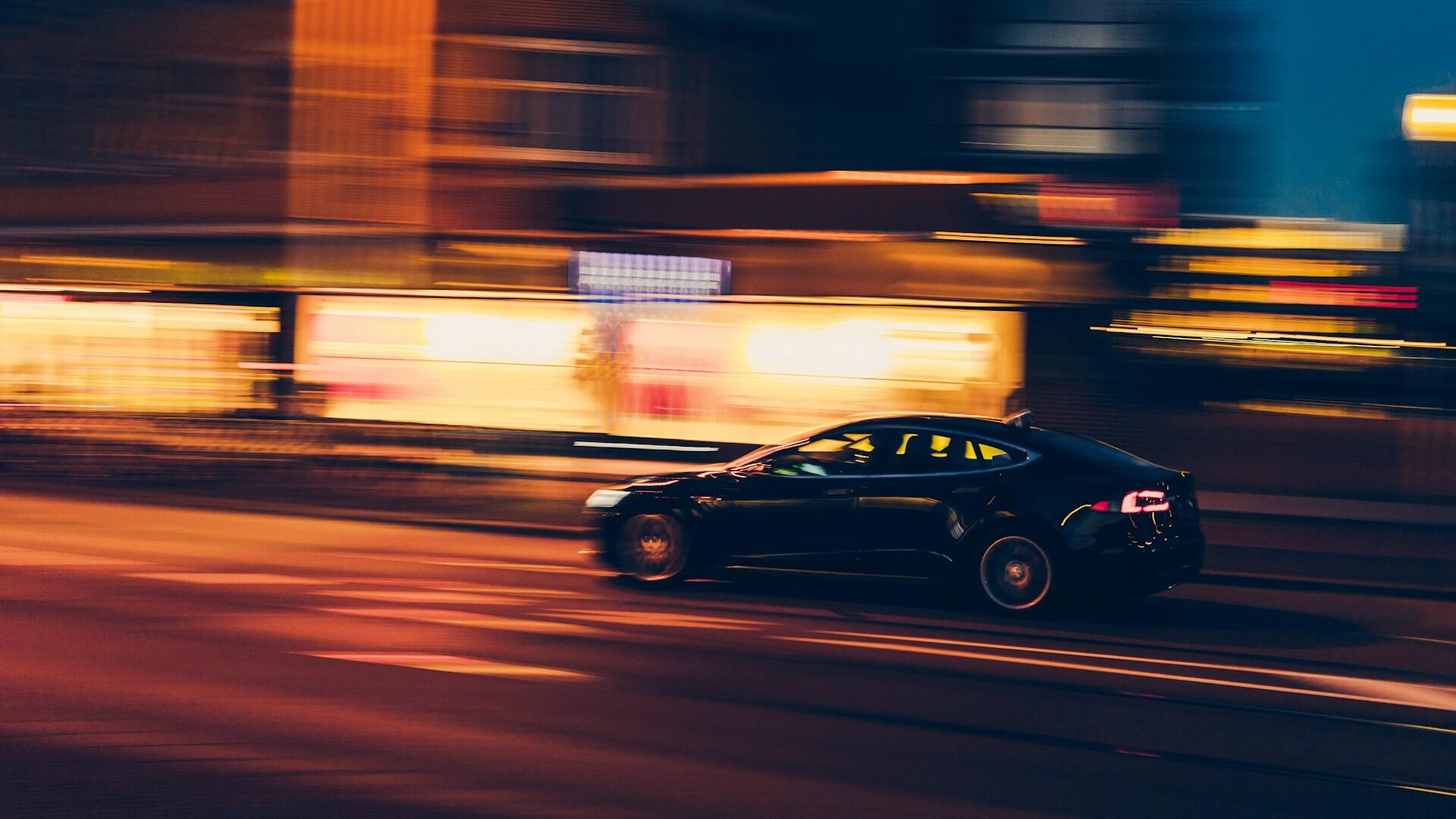 A sleek black car speeds through a colorful city street at night, with vibrant lights creating a dynamic blur around it.