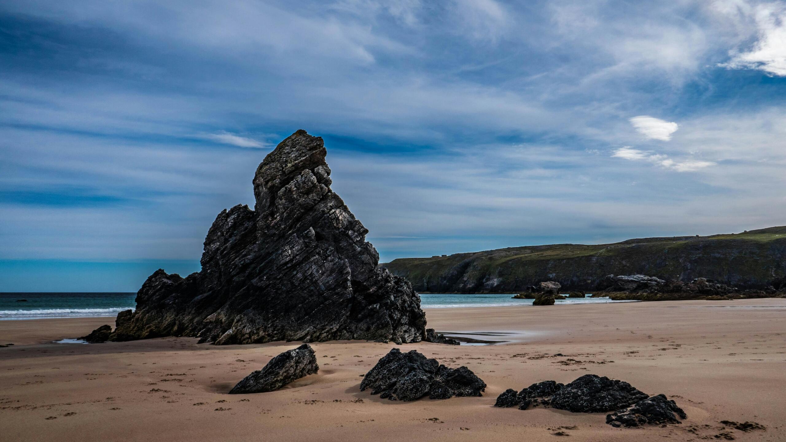 a rock formation on a beach. The sky is cloudy and a forest can be seen in the background.