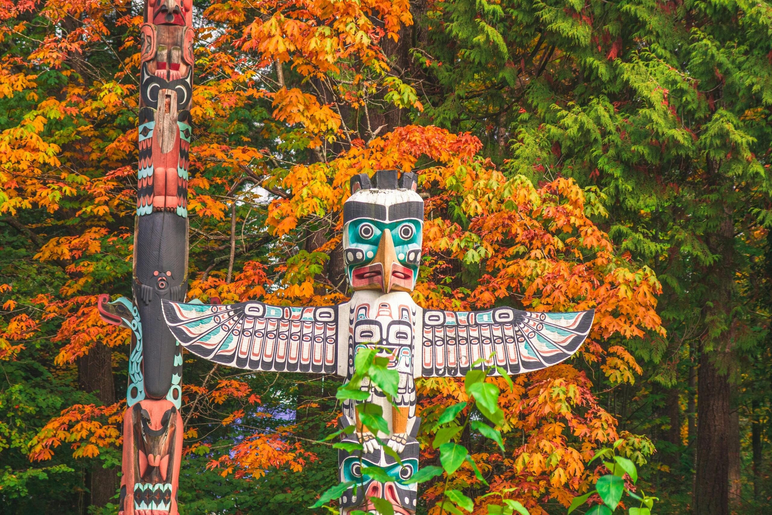 two brightly painted totem poles in front of trees. One tree has orange leaves.