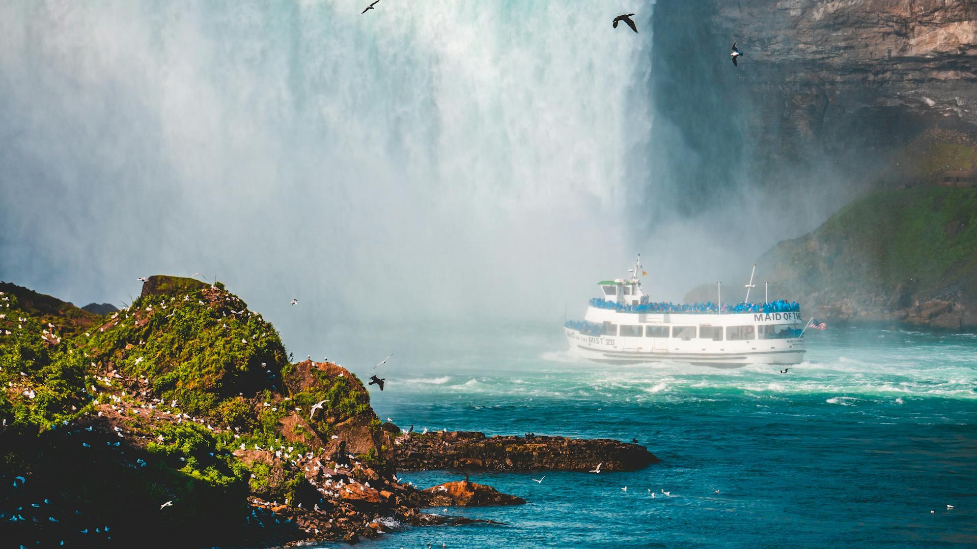 A boat, "Maid of the Mist," navigates the mist near Niagara Falls, surrounded by green cliffs and birds in flight.