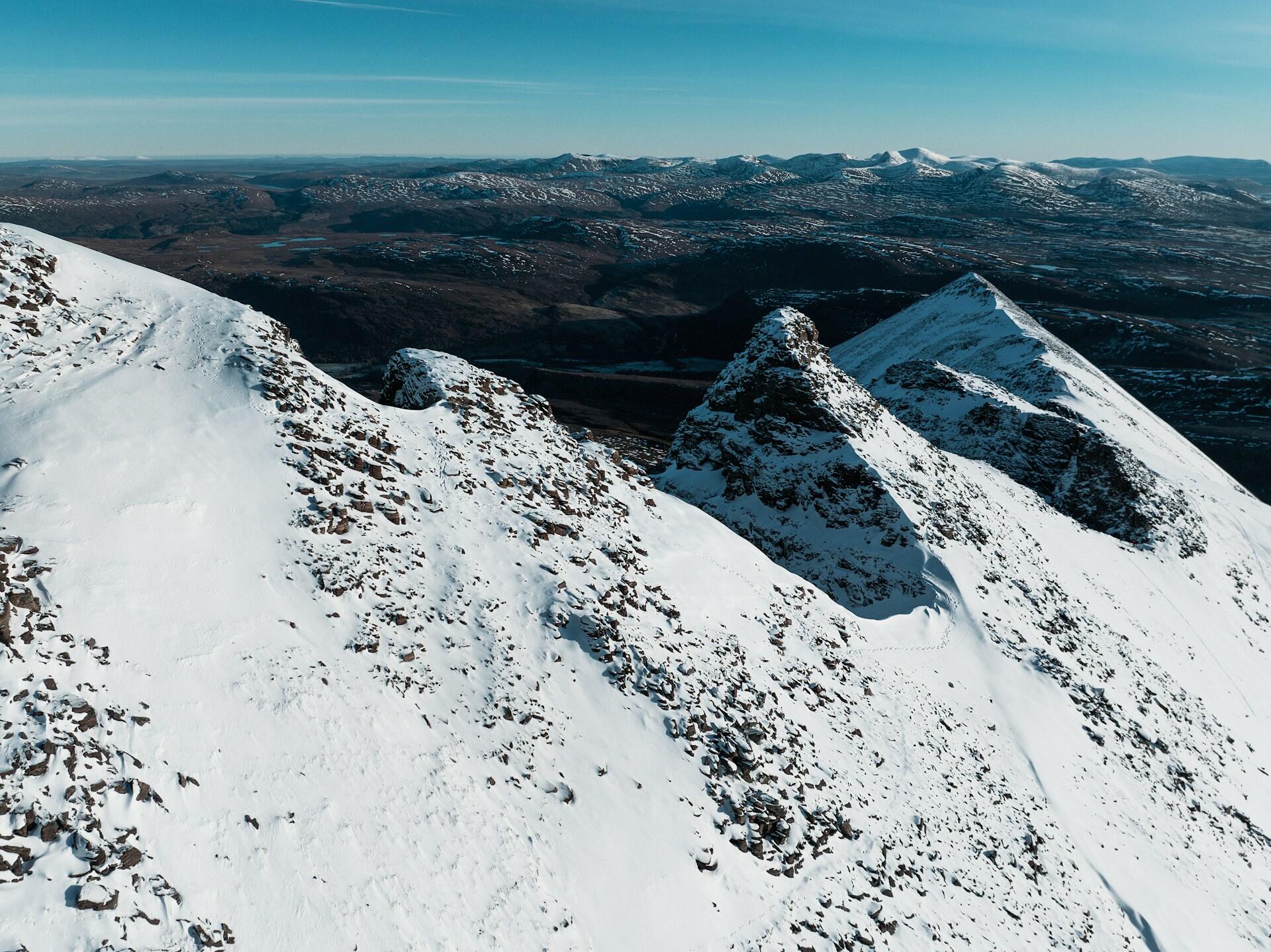 Snow-covered mountain peaks rise against a clear blue sky, overlooking a vast, rugged landscape dotted with patches of snow.