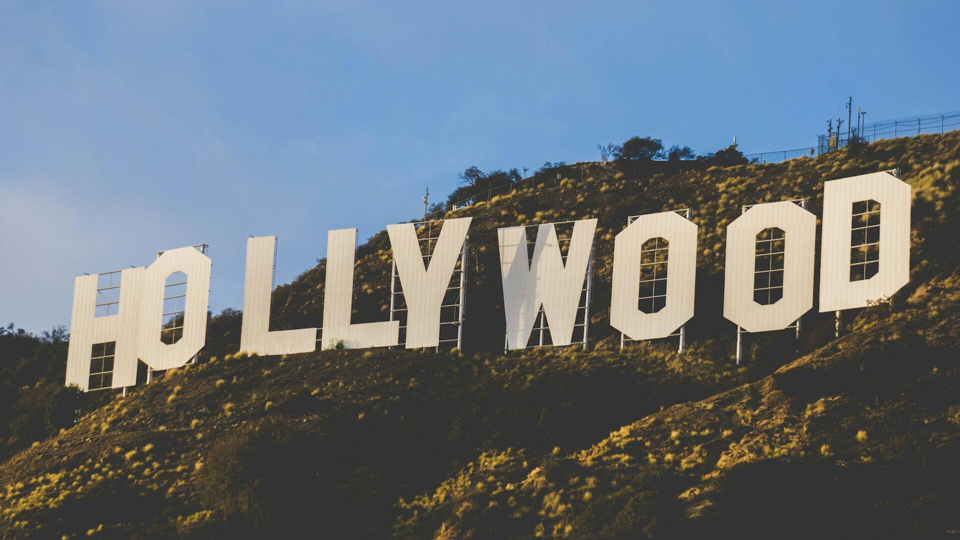 The iconic Hollywood sign, partially obscured, stands on a hillside under a blue sky with scattered clouds.