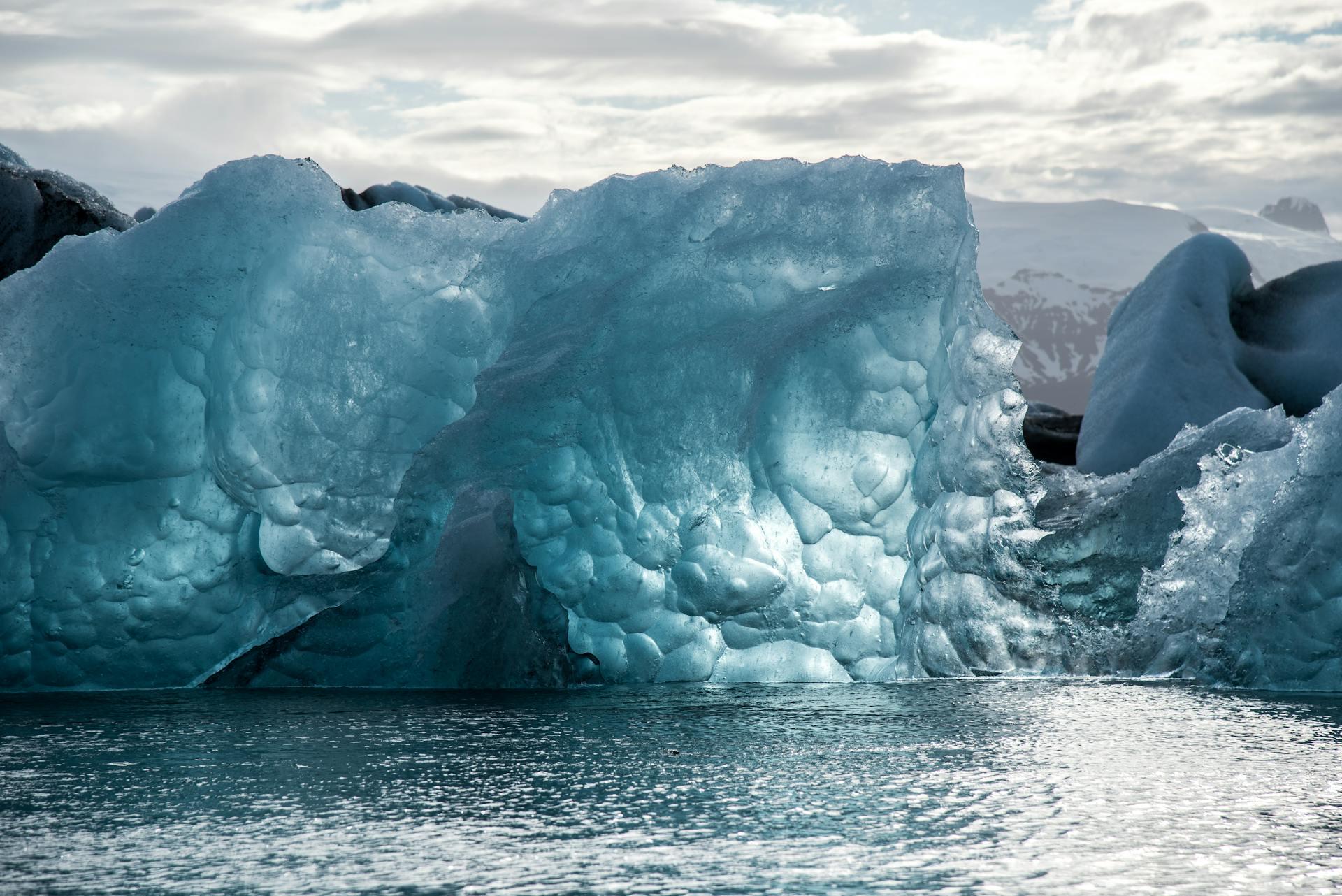 A close-up of a translucent blue ice sculpture, partially submerged in water, with soft clouds and mountains in the background.