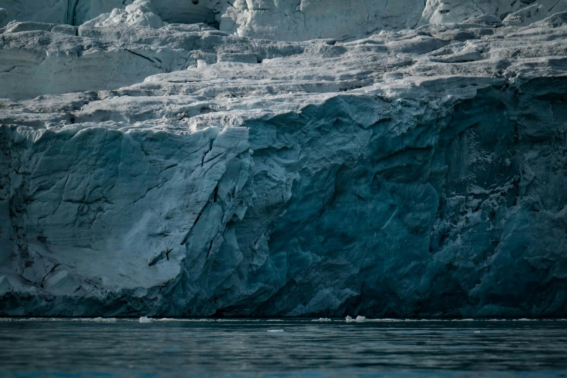 A close-up of a textured ice cliff with deep blue tones, reflecting in the calm water below, highlighting glacial formations.