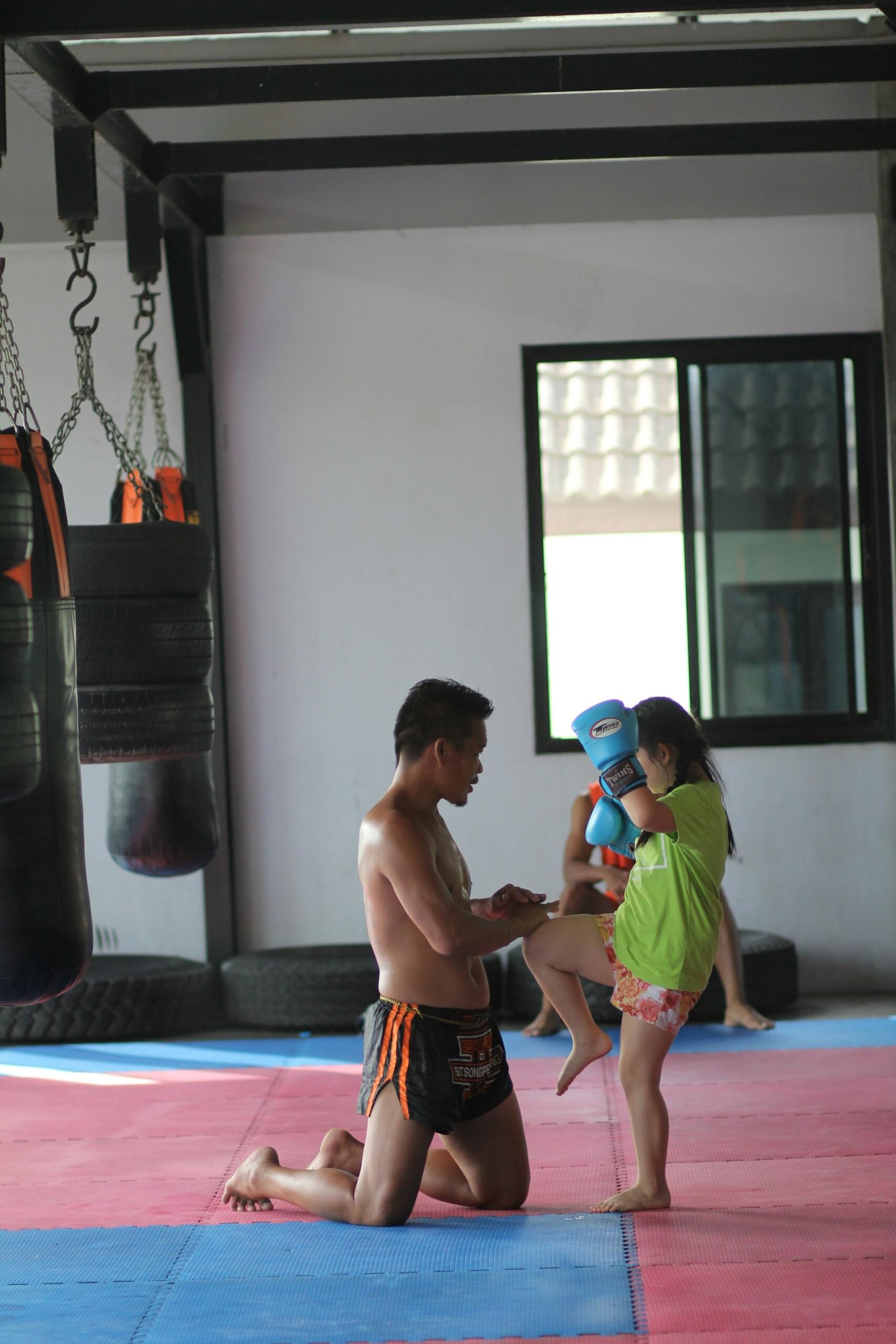 A trainer kneels on a mat while instructing a young girl in a bright green shirt, who practices a kick while wearing boxing gloves.