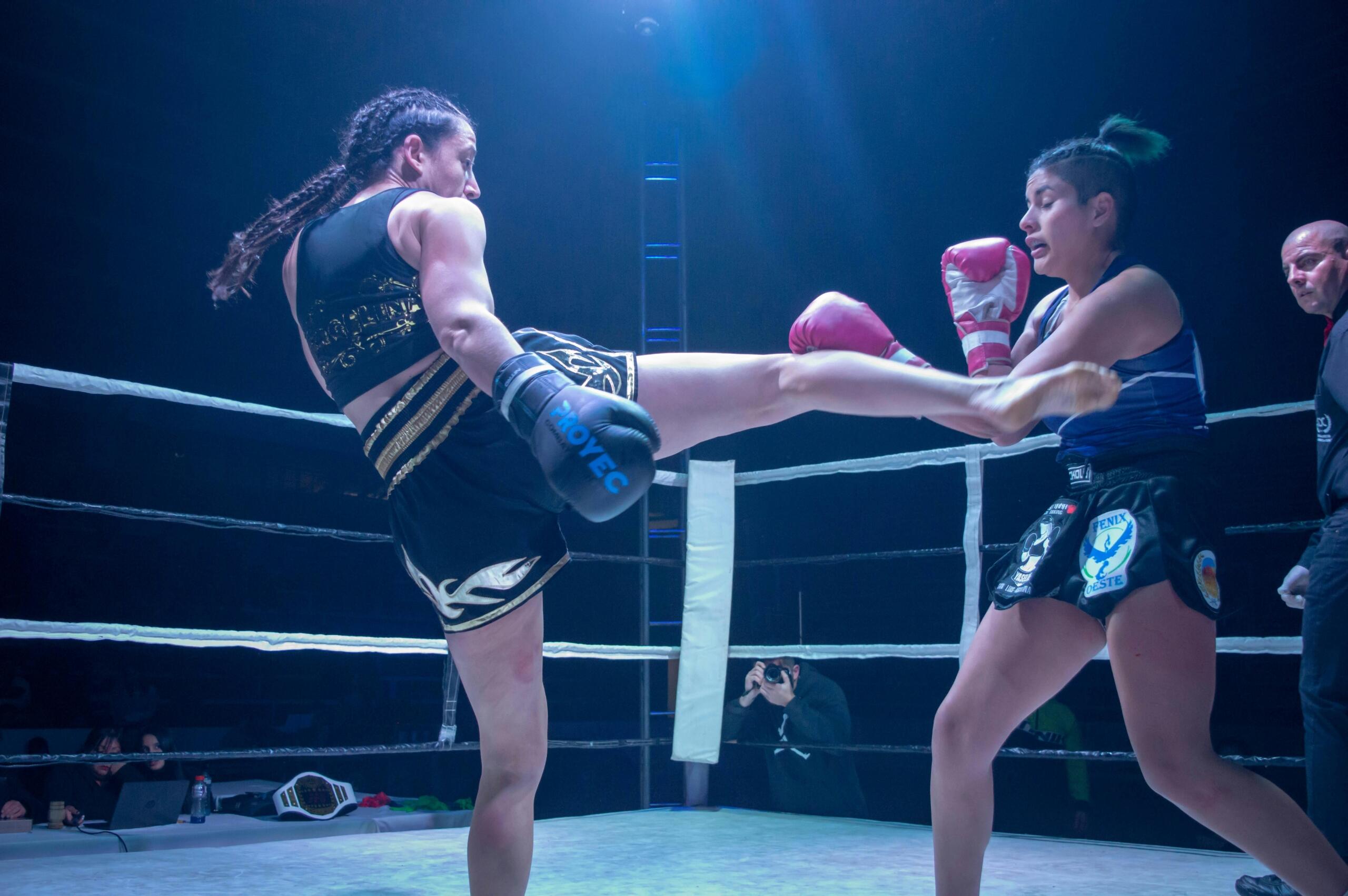 Two female fighters exchanging strikes in a kickboxing match inside a boxing ring, with one delivering a high kick while the referee watches closely.