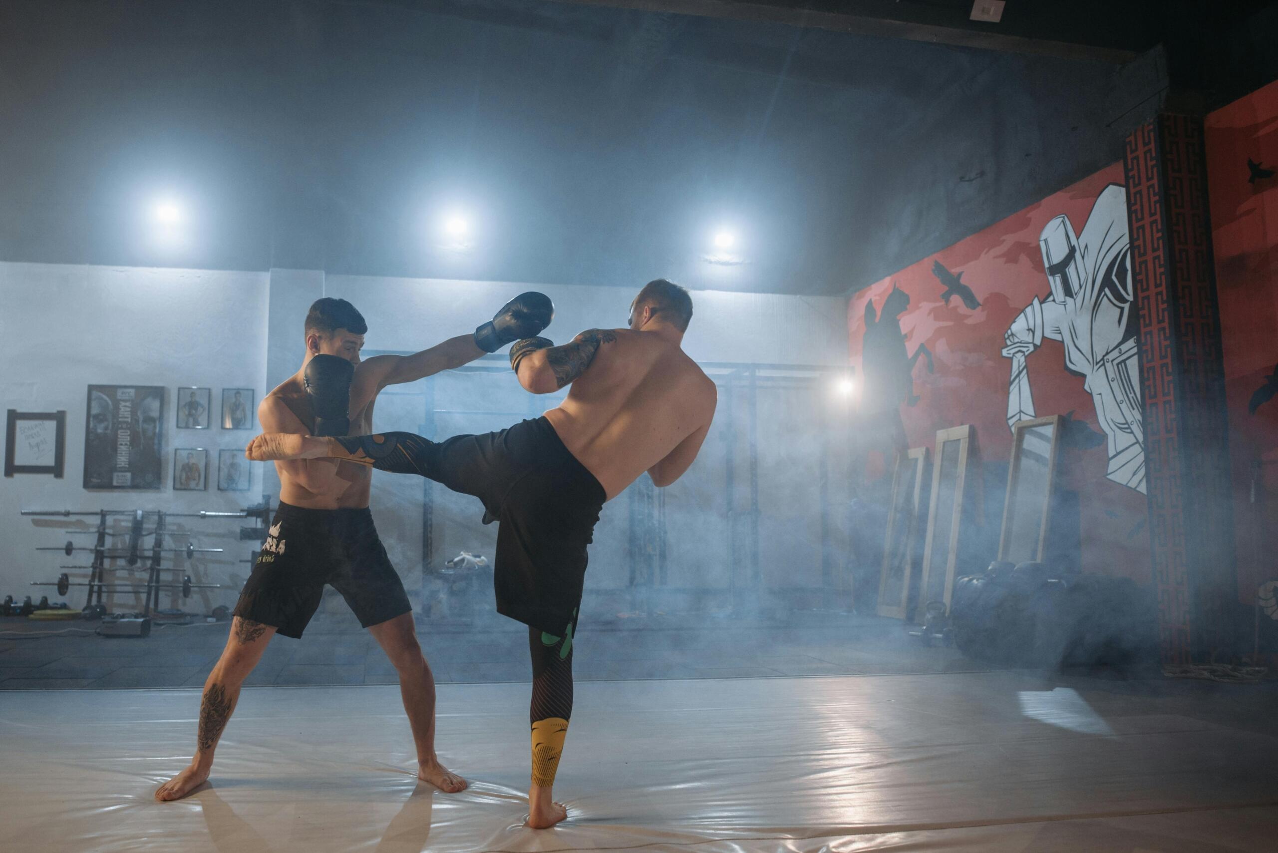Two fighters sparring in an MMA gym, one delivering a kick while the other extends a punch. The gym has a misty atmosphere with bright overhead lights and a mural of a medieval knight on the wall. Various training equipment, including weight racks and framed pictures, are visible in the background.