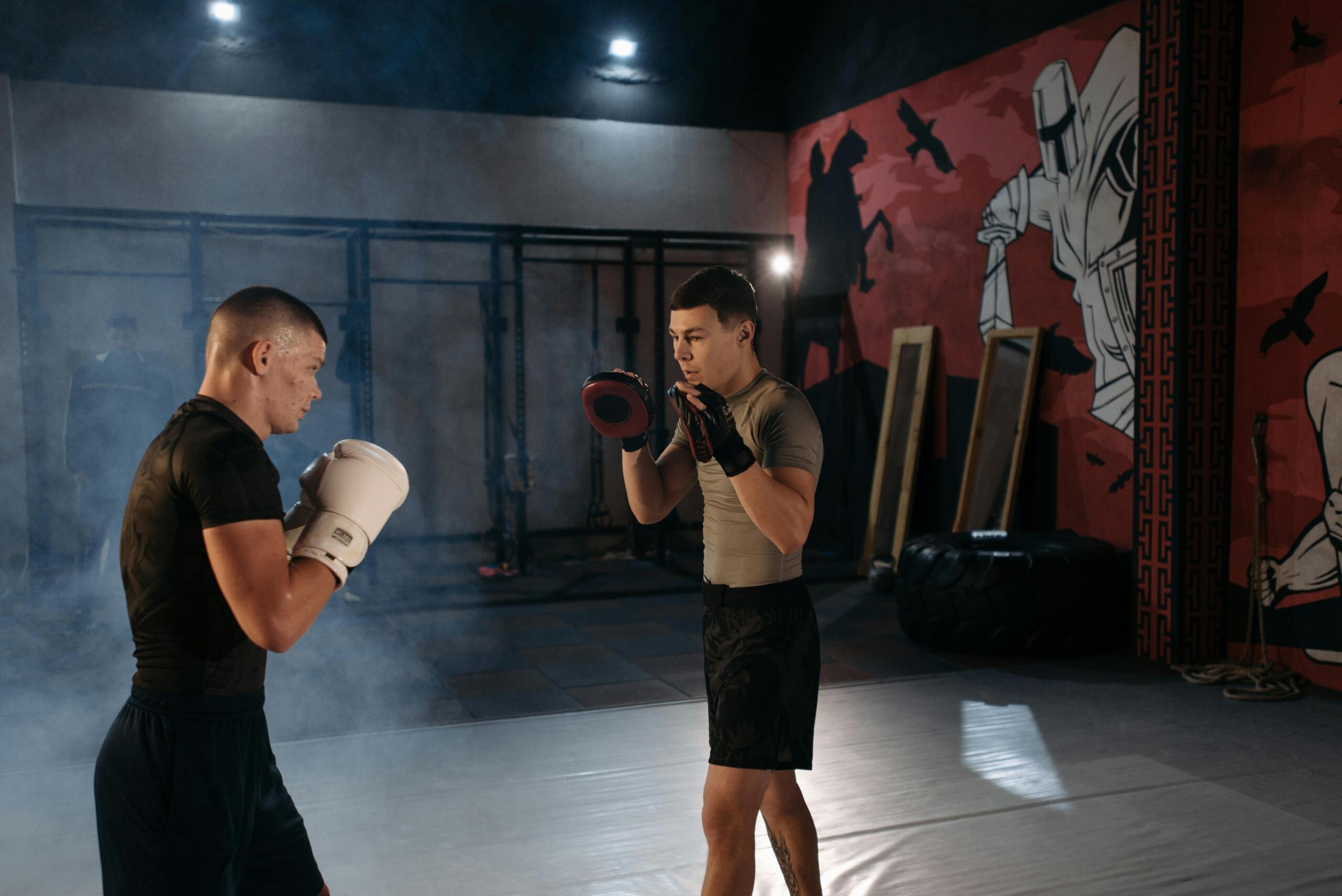 Two men training in a dimly lit gym with a red and black mural on the wall. One man, wearing a black shirt, dark shorts, and white boxing gloves, is in a fighting stance. The other man, dressed in a gray shirt and black shorts, holds red focus mitts, preparing for a training drill. Gym equipment, including tires and wooden boards, is visible in the background.