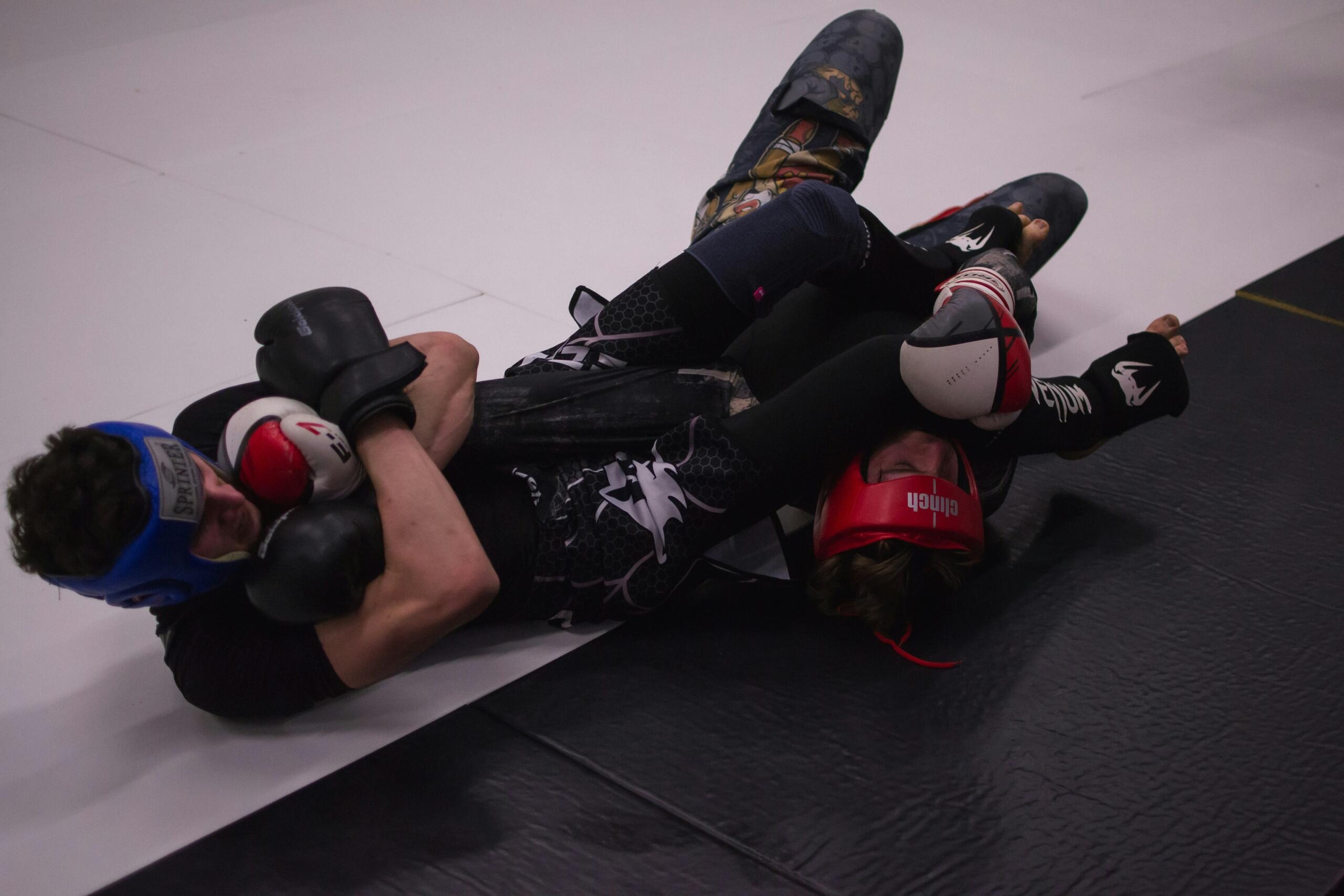 Two MMA fighters sparring on a mat, both wearing protective gear. One fighter, in a red headguard and black gloves, has their opponent locked in an armbar submission hold. The other fighter, wearing a blue headguard and black gloves, is on the ground attempting to defend against the hold. The background consists of a white and black mat.