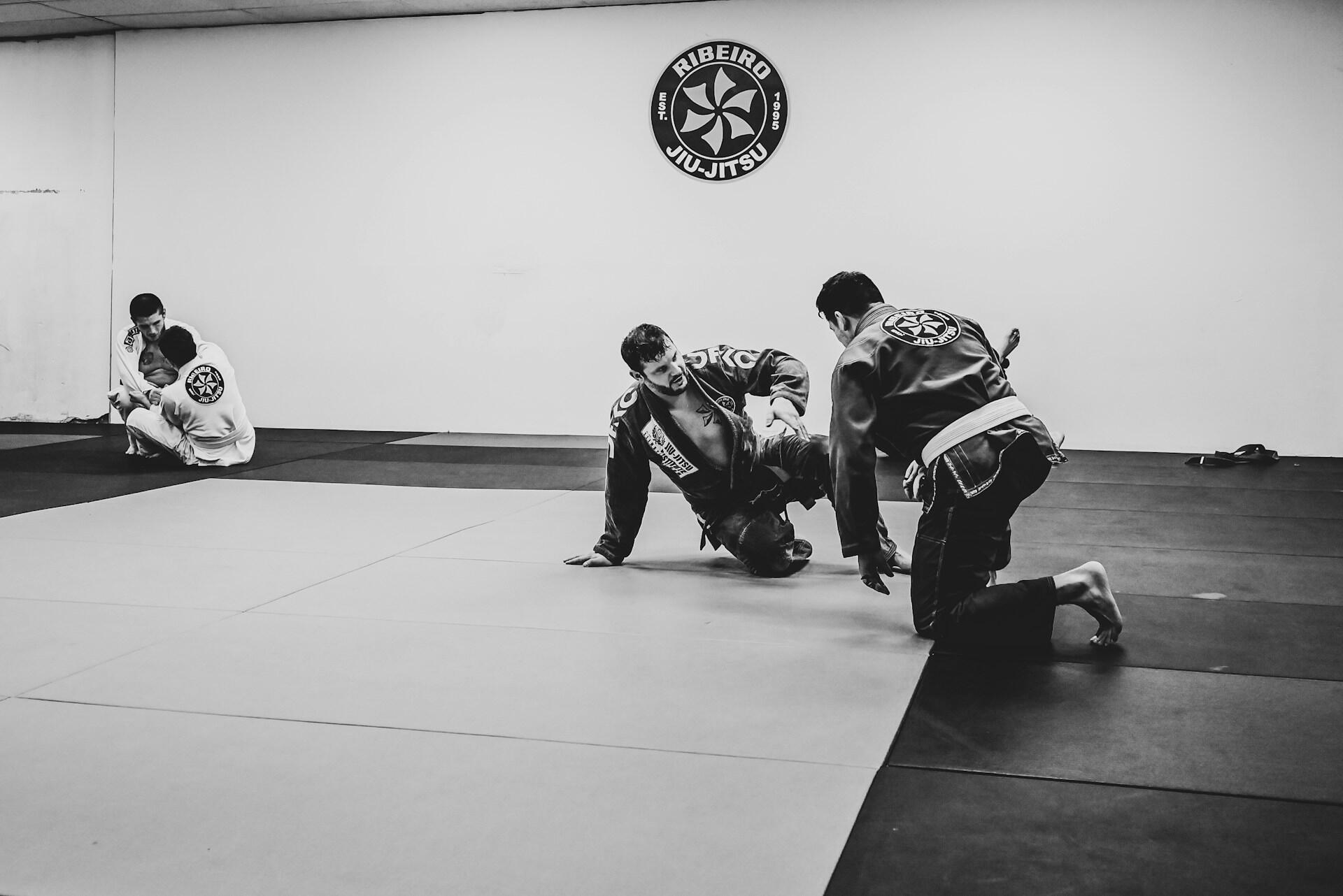Two Brazilian Jiu-Jitsu practitioners grapple on mats, while a third sits quietly in the background, showcasing an active training environment.
