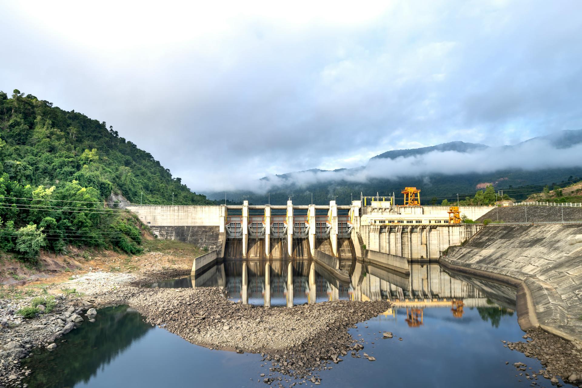 A dam with a series of gates, surrounded by lush green mountains and reflected in calm water, under a partially cloudy sky.