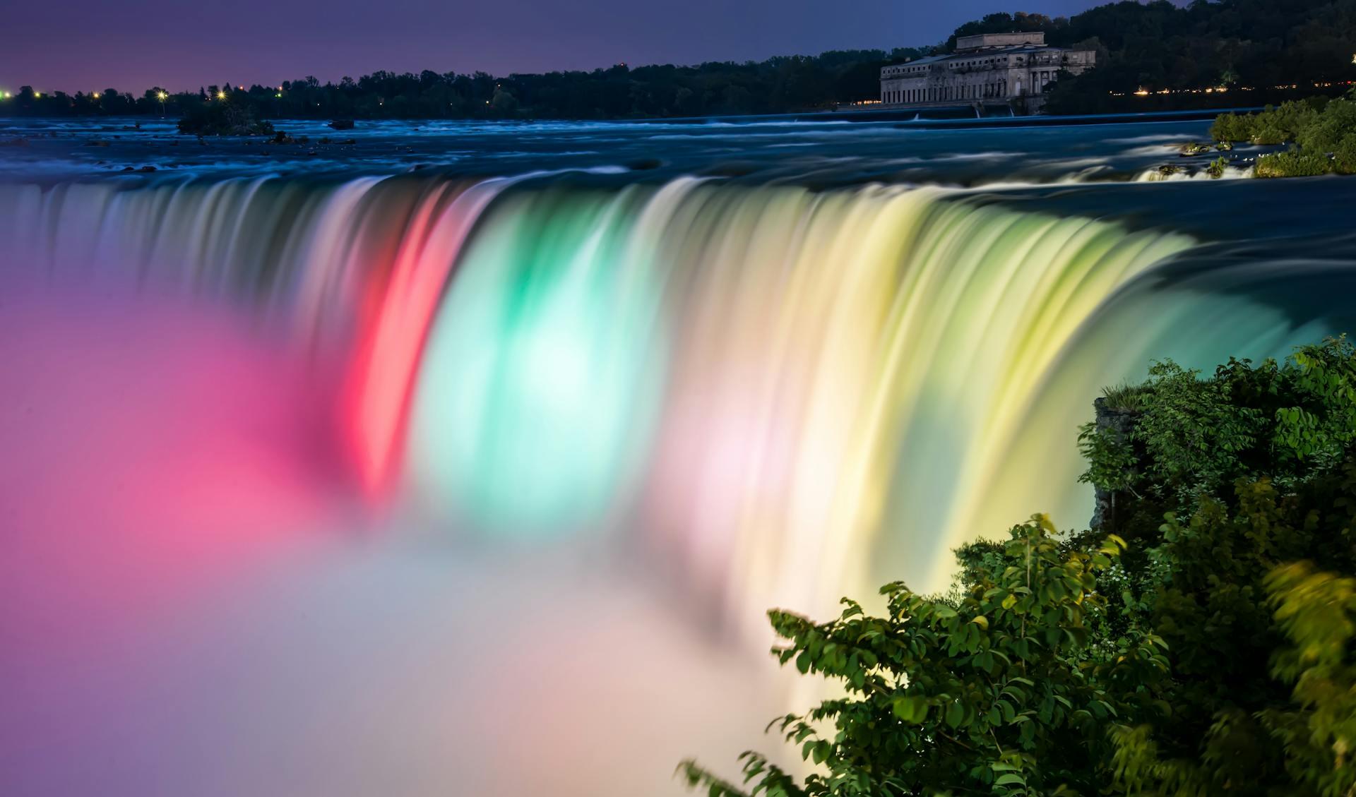 Niagara Falls illuminated in vibrant colors under a twilight sky, with lush greenery in the foreground and soft water flow effects.