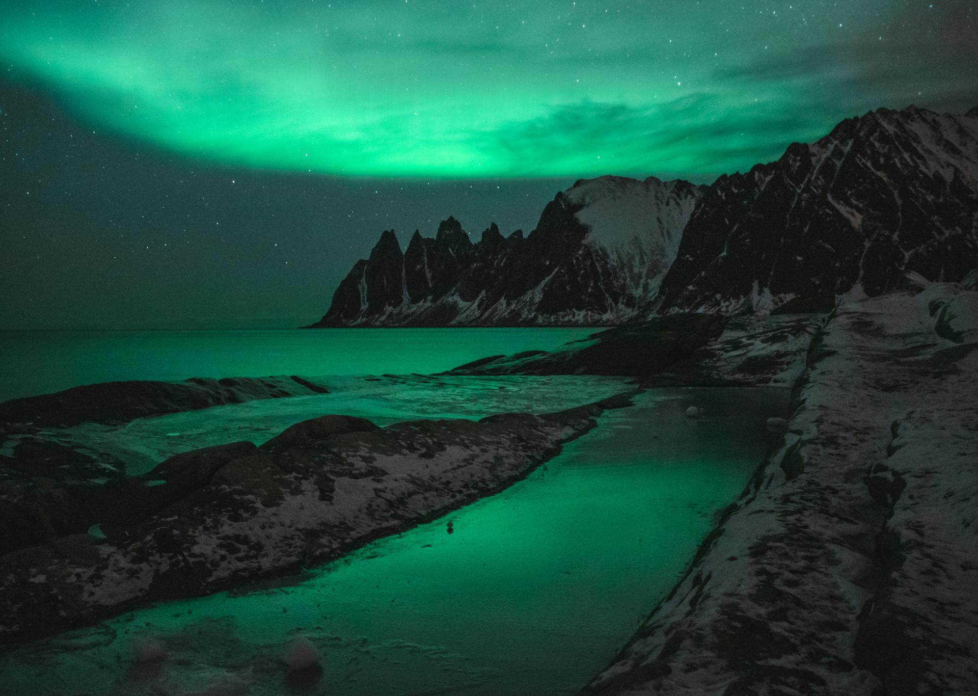 Majestic green auroras dance above jagged, snow-capped mountains, reflecting on a tranquil water surface under a starry night sky.
