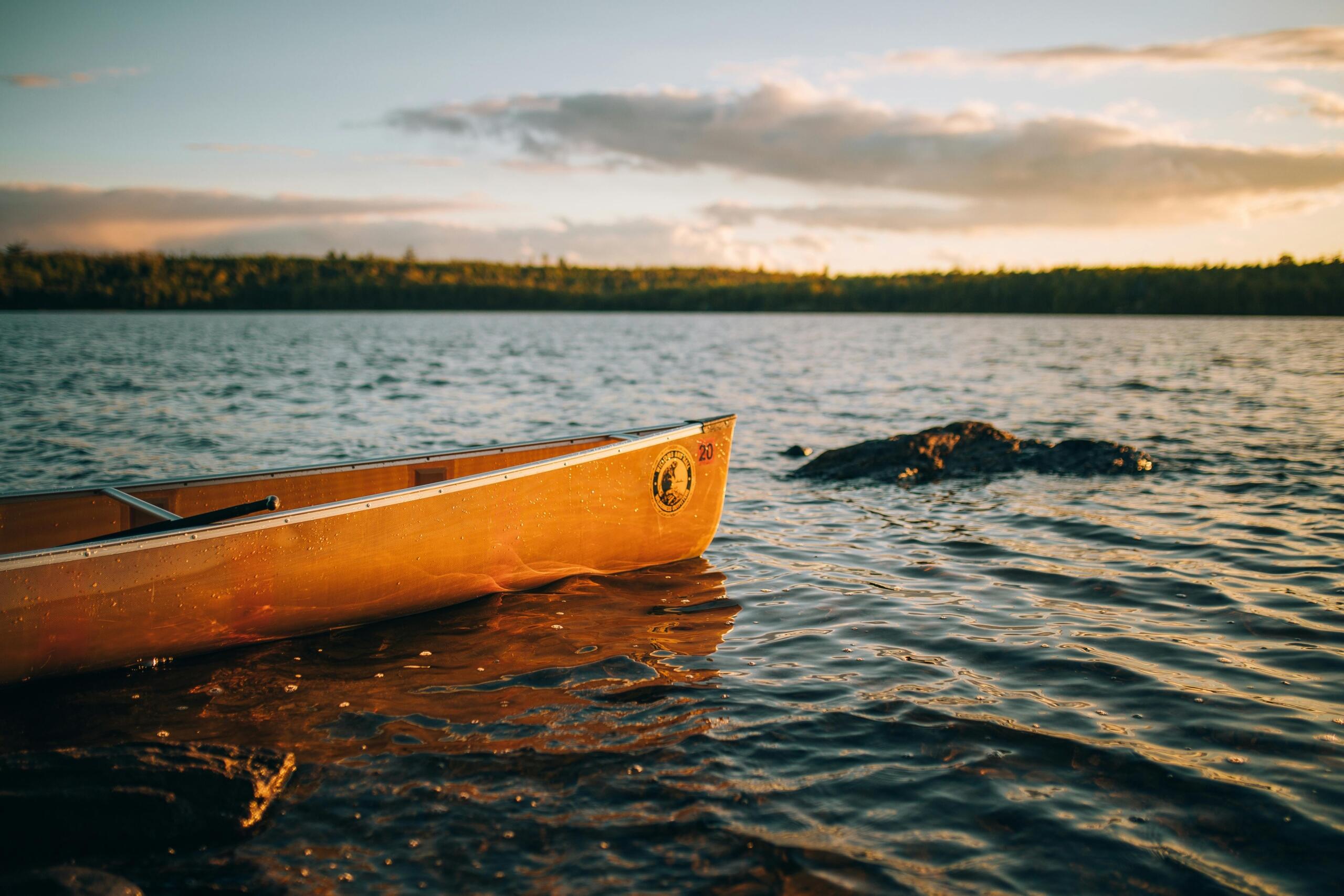 an empty wooden canoe sits on a rocky shore. Forests can be seen across the water.