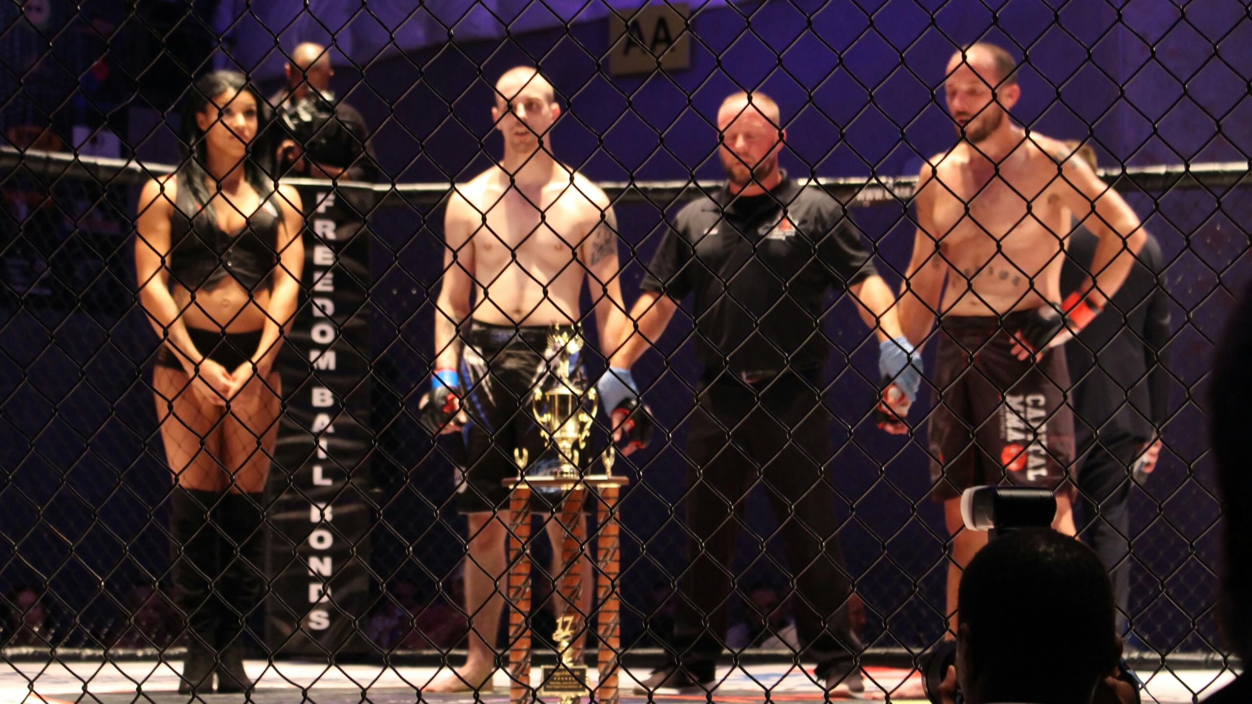 Two shirtless MMA fighters stand inside a caged octagon, wearing blue and red gloves. A referee in black stands between them. A woman in black attire with long boots stands nearby. A trophy is placed in the center. The cage fence is visible in the foreground, and a photographer captures the moment. A sign with “AA” hangs in the dimly lit background.