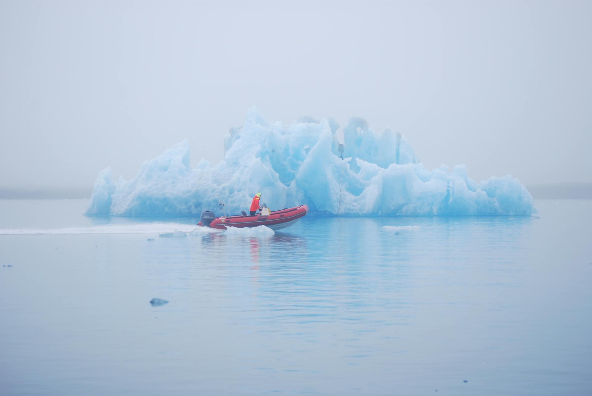 A small red boat navigates icy waters, passing a large blue iceberg, under a foggy, overcast sky.