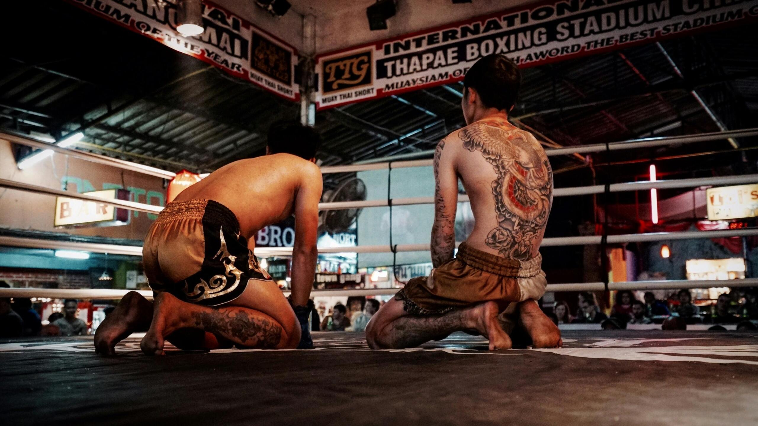 Two Muay Thai fighters kneeling in a traditional pre-fight ritual inside a boxing stadium, surrounded by spectators and gym banners.
