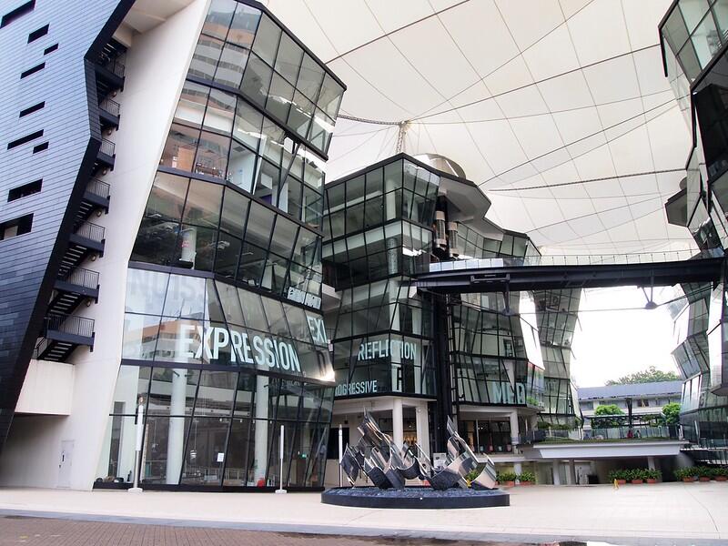 A modern architectural building with large glass windows reflecting the surroundings. The structure features bold, angled designs with words like “EXPRESSION” and “REFLECTION” displayed on the glass. A canopy covers the open courtyard, and a pedestrian bridge connects sections of the building. A sculptural installation sits in the center of the courtyard.