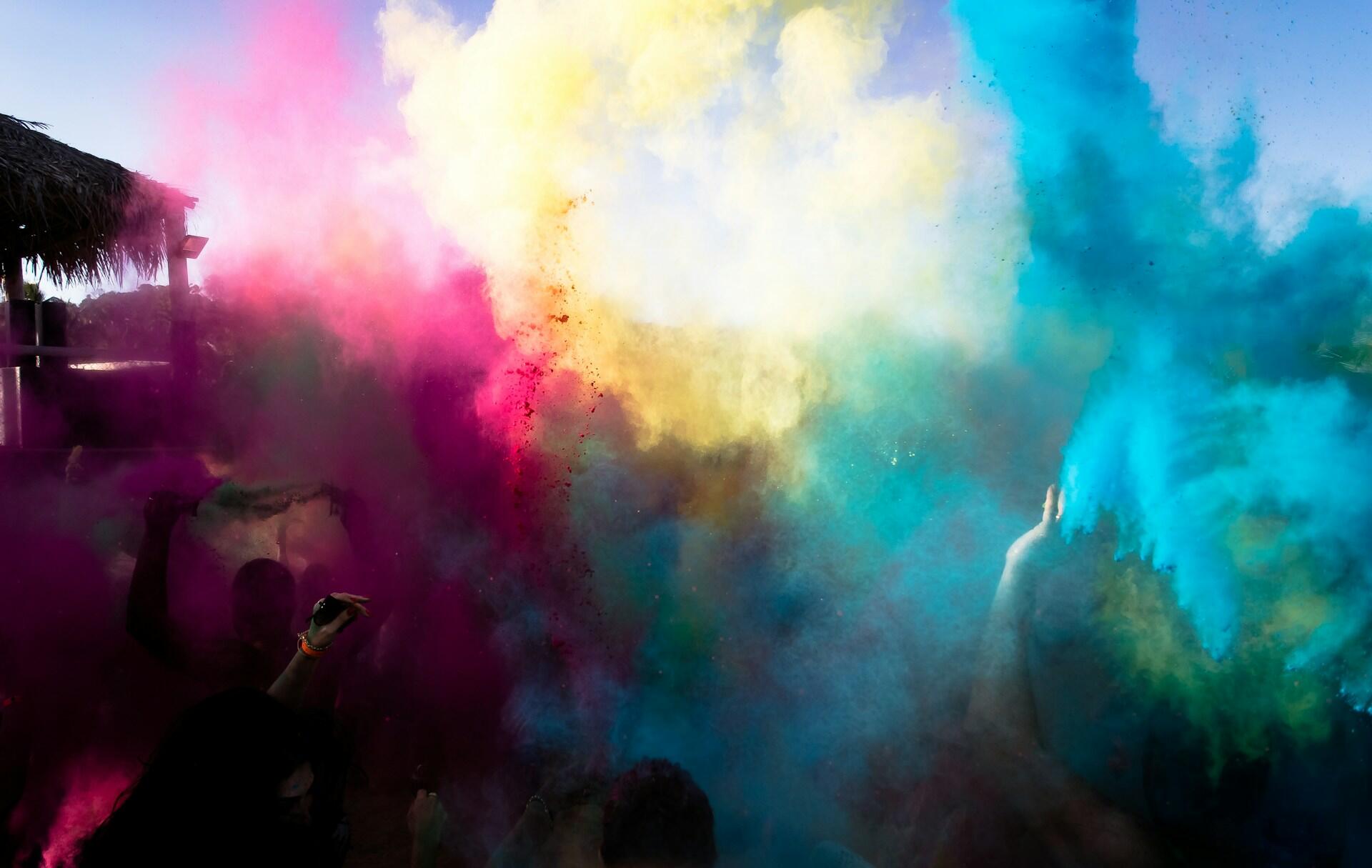 A vibrant cloud of colored powders in pink, yellow, and blue envelops a festive outdoor scene, capturing the essence of celebration.